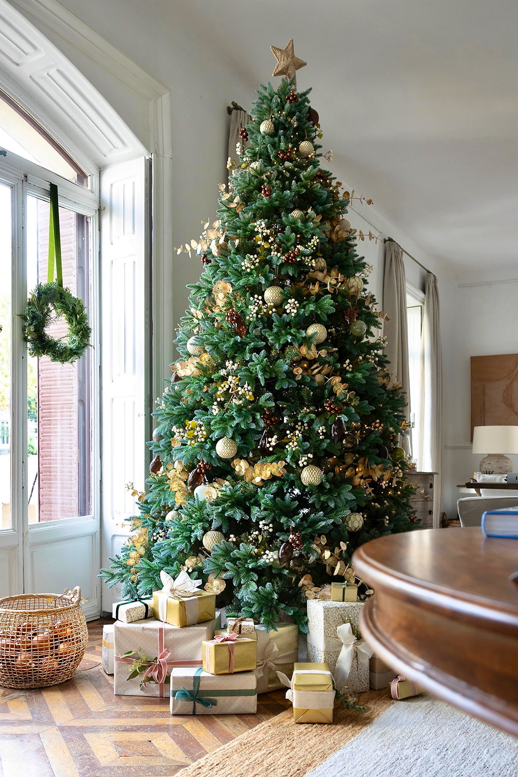 Árbol de Navidad decorado elegantemente con adornos dorados y blancos, rodeado de regalos envueltos en papel kraft y dorado, junto a una ventana alta con una corona navideña.