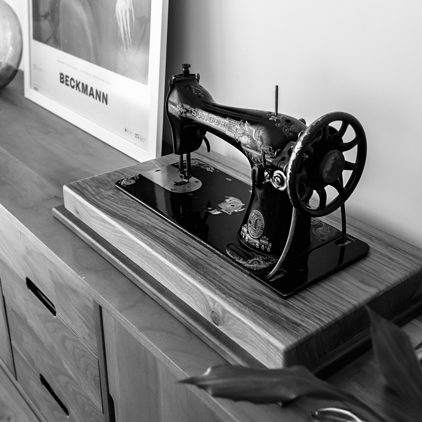 Máquina de coser vintage en blanco y negro, con detalles ornamentales, situada sobre un mueble de madera con un póster de Beckmann al fondo