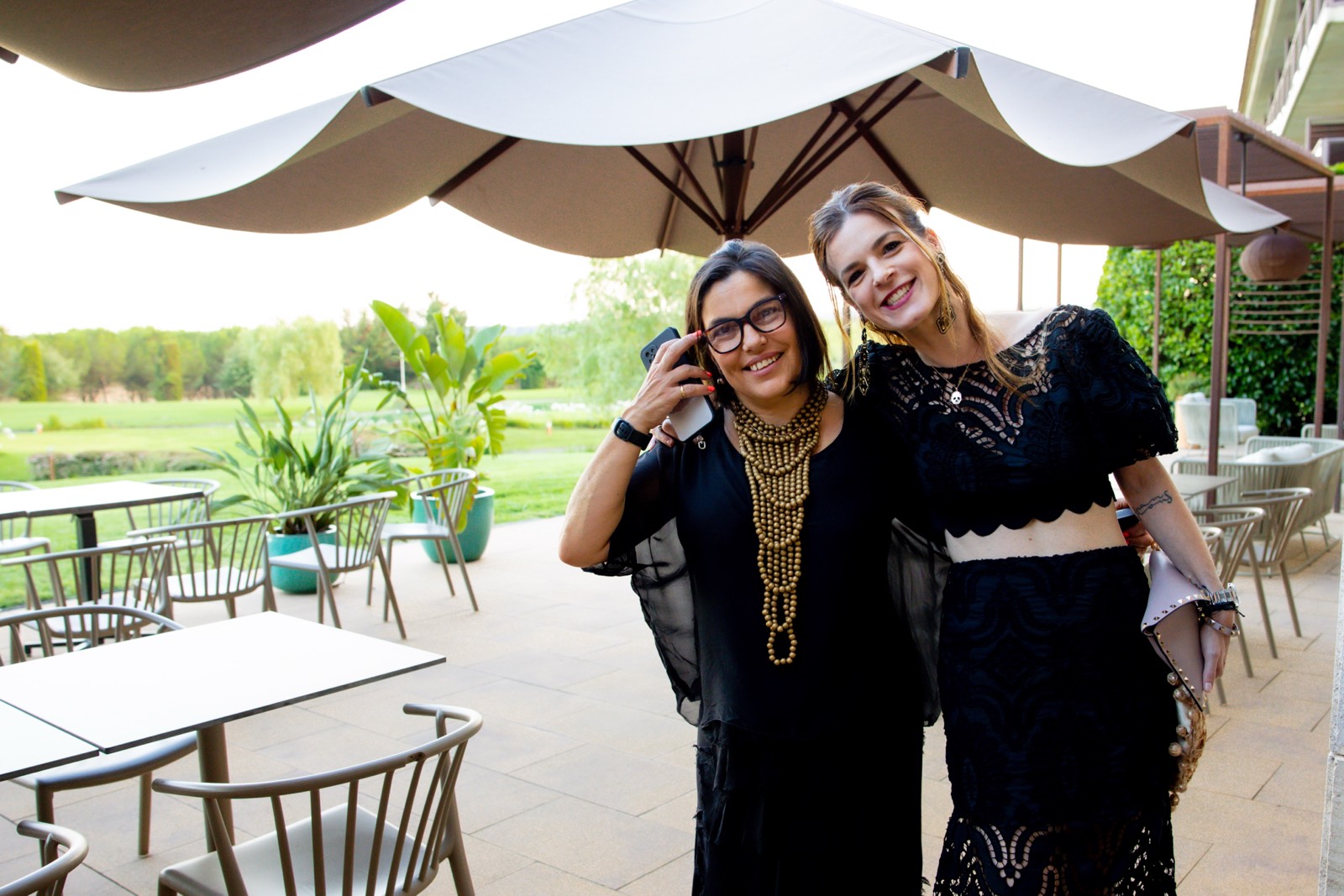 Dos mujeres sonrientes vestidas de negro, con un paisaje verde y una terraza con sombrilla en el fondo