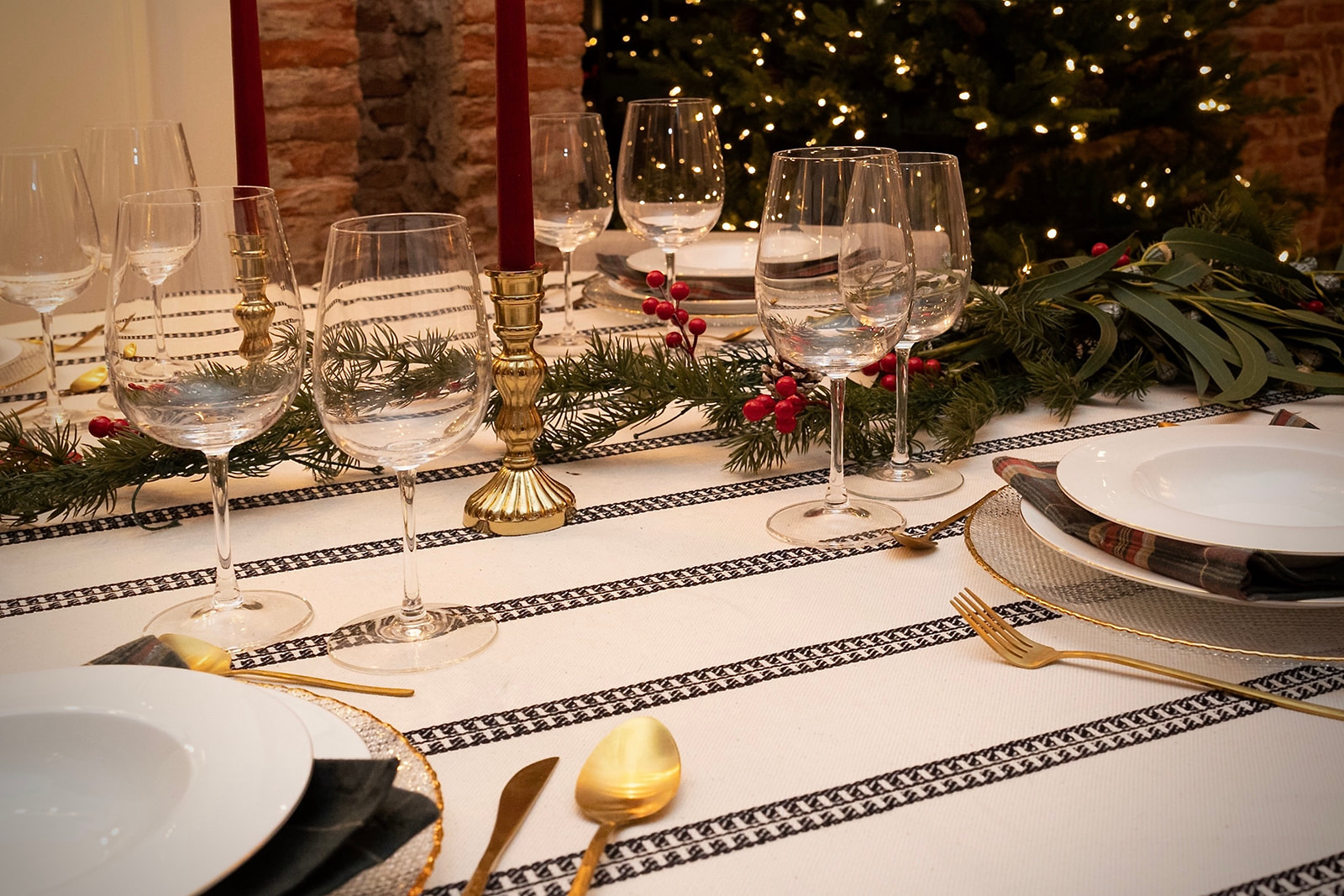 Mesa elegante decorada para Navidad con mantel blanco, copas de vino, platos, cubiertos dorados, velas rojas y adornos verdes. Al fondo se ve un árbol de Navidad iluminado.