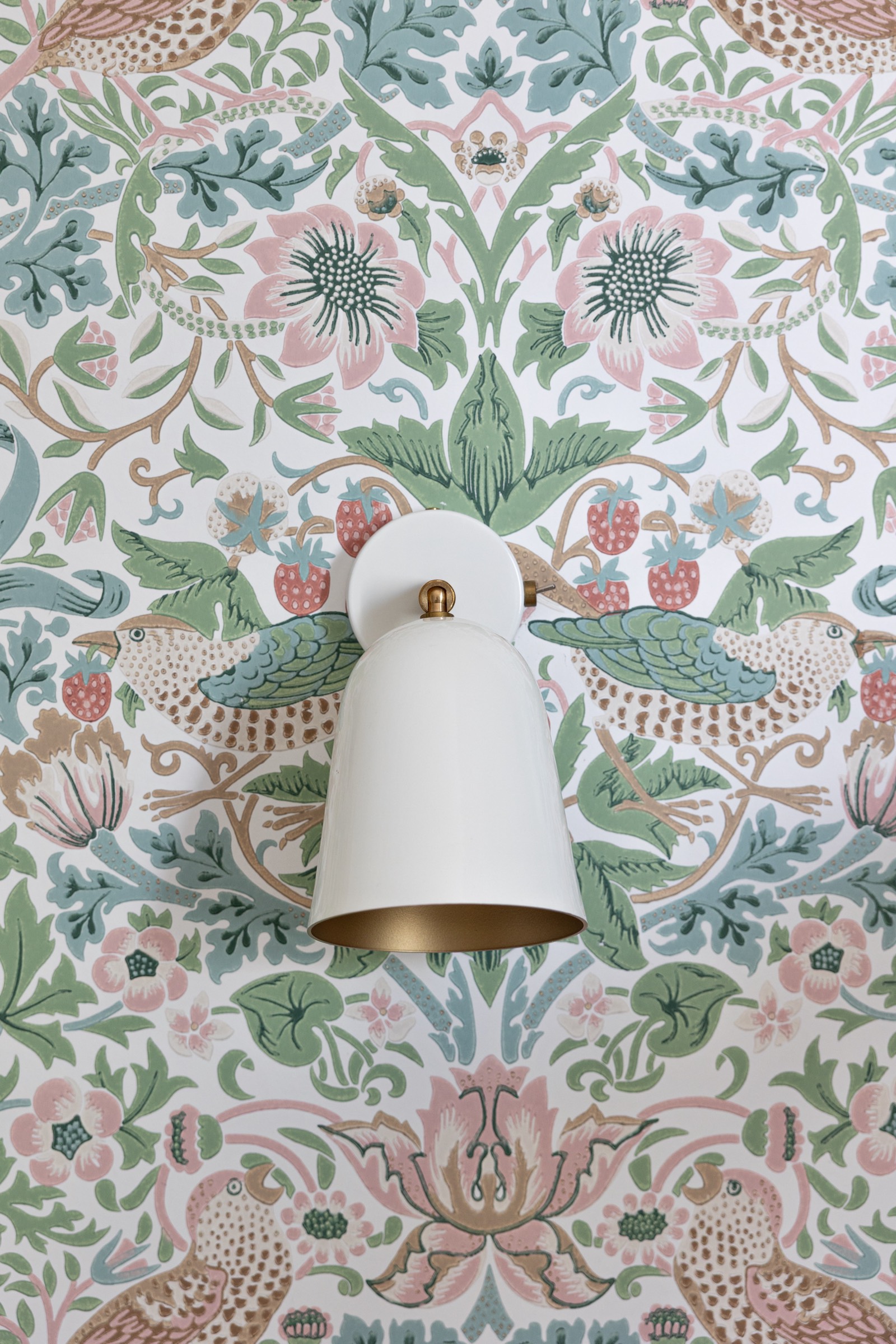 Lámpara de pared blanca con interior dorado montada sobre un papel tapiz intrincado con diseño de pájaros, flores y fresas en tonos pastel