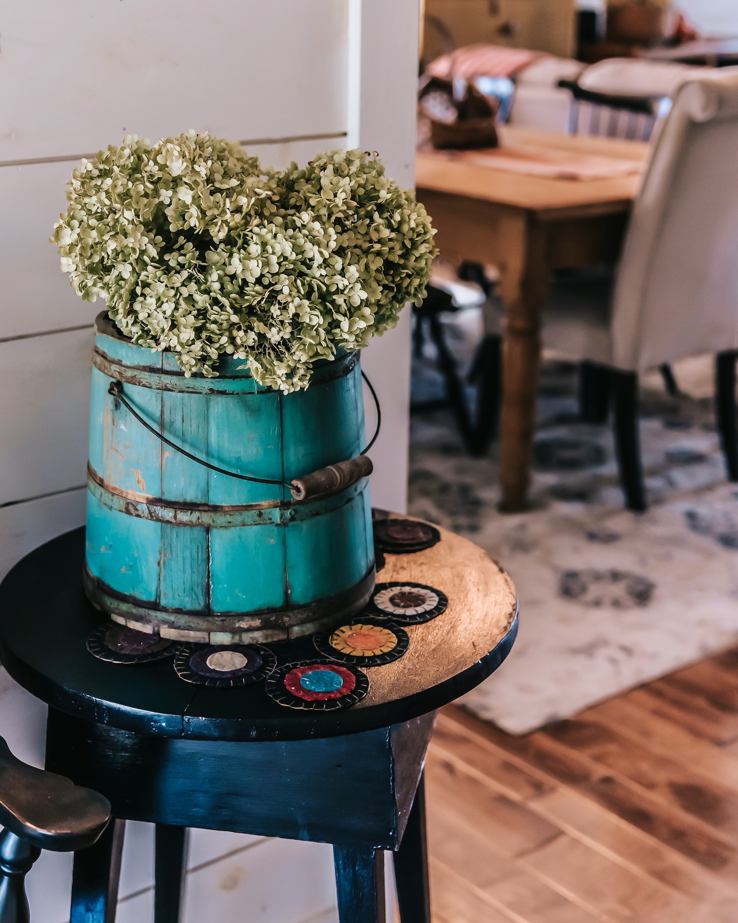 Arreglo de hortensias blancas en un cubo vintage de color turquesa desgastado, colocado sobre una mesa auxiliar azul oscuro decorada con círculos ornamentales. Decoración de estilo rústico y campestre.