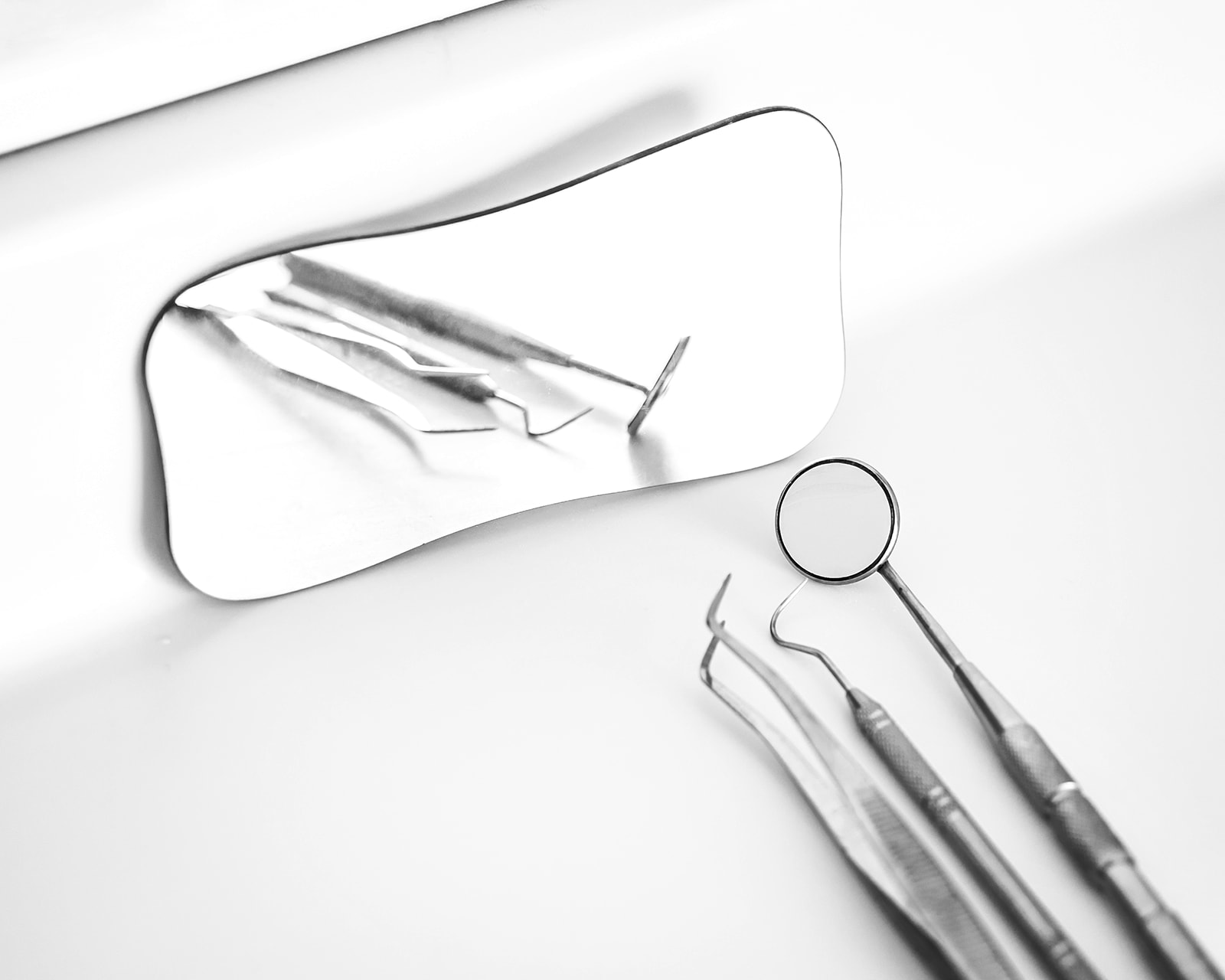Imagen en blanco y negro de instrumentos dentales, incluyendo un espejo dental y herramientas de exploración, junto a un espejo rectangular más grande que refleja los instrumentos.