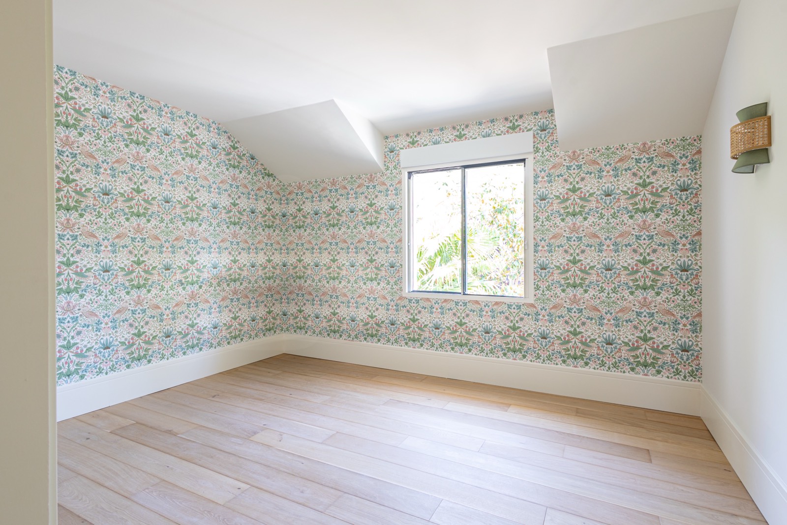 Habitación vacía con paredes decoradas con papel tapiz floral en tonos pastel, piso de madera clara y ventana con vista a plantas