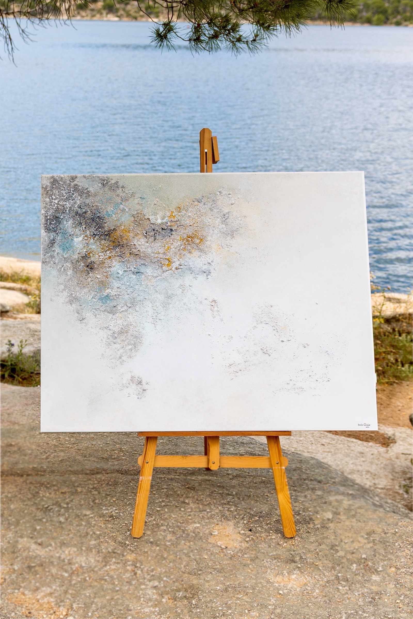 Lienzo blanco con pintura abstracta en tonos grises, azules y dorados sobre un caballete de madera junto a un lago tranquilo. Ramas de pino enmarcan la parte superior de la imagen.