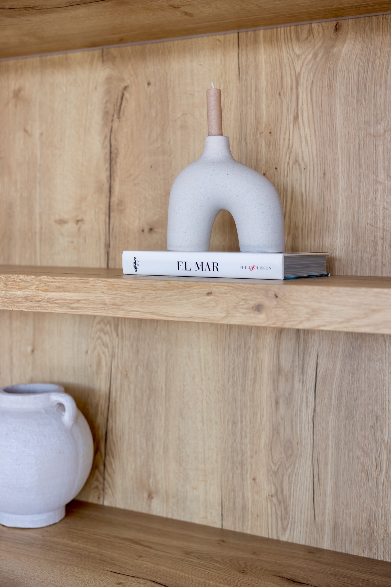 Estantería de madera con un candelabro moderno en forma de arco sobre un libro titulado 'El Mar'. En el estante inferior se ve una vasija blanca.