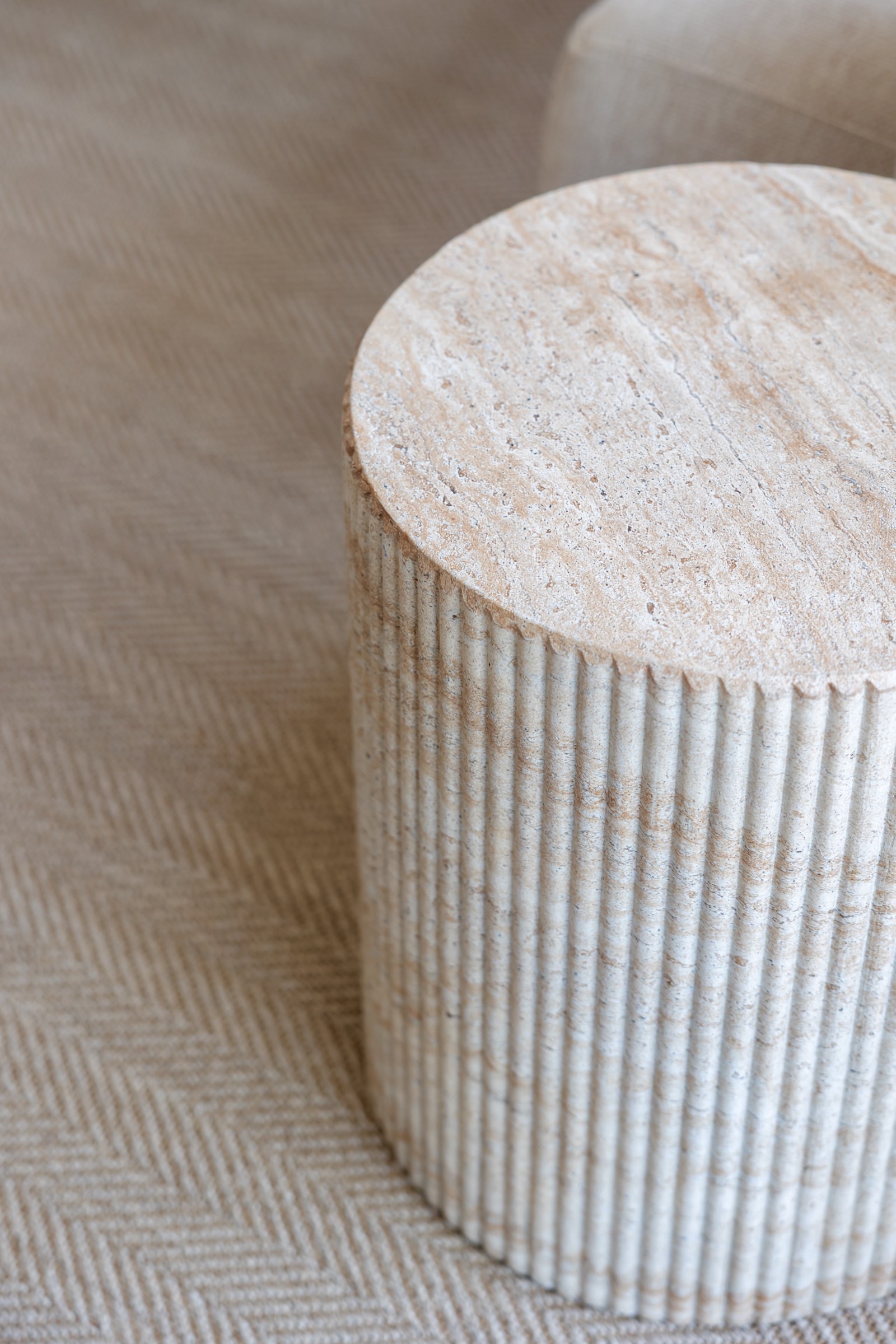 Primer plano de una mesa auxiliar cilíndrica de travertino con superficie lisa y lados acanalados verticalmente, colocada sobre una tela con patrón de espiga.