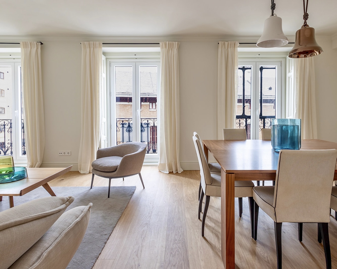 Salón-comedor luminoso con estilo moderno, ventanales grandes, cortinas blancas, mesa de madera, sillas tapizadas, sillones cómodos y lámparas colgantes. El espacio tiene un aspecto acogedor y elegante con tonos claros y neutros.