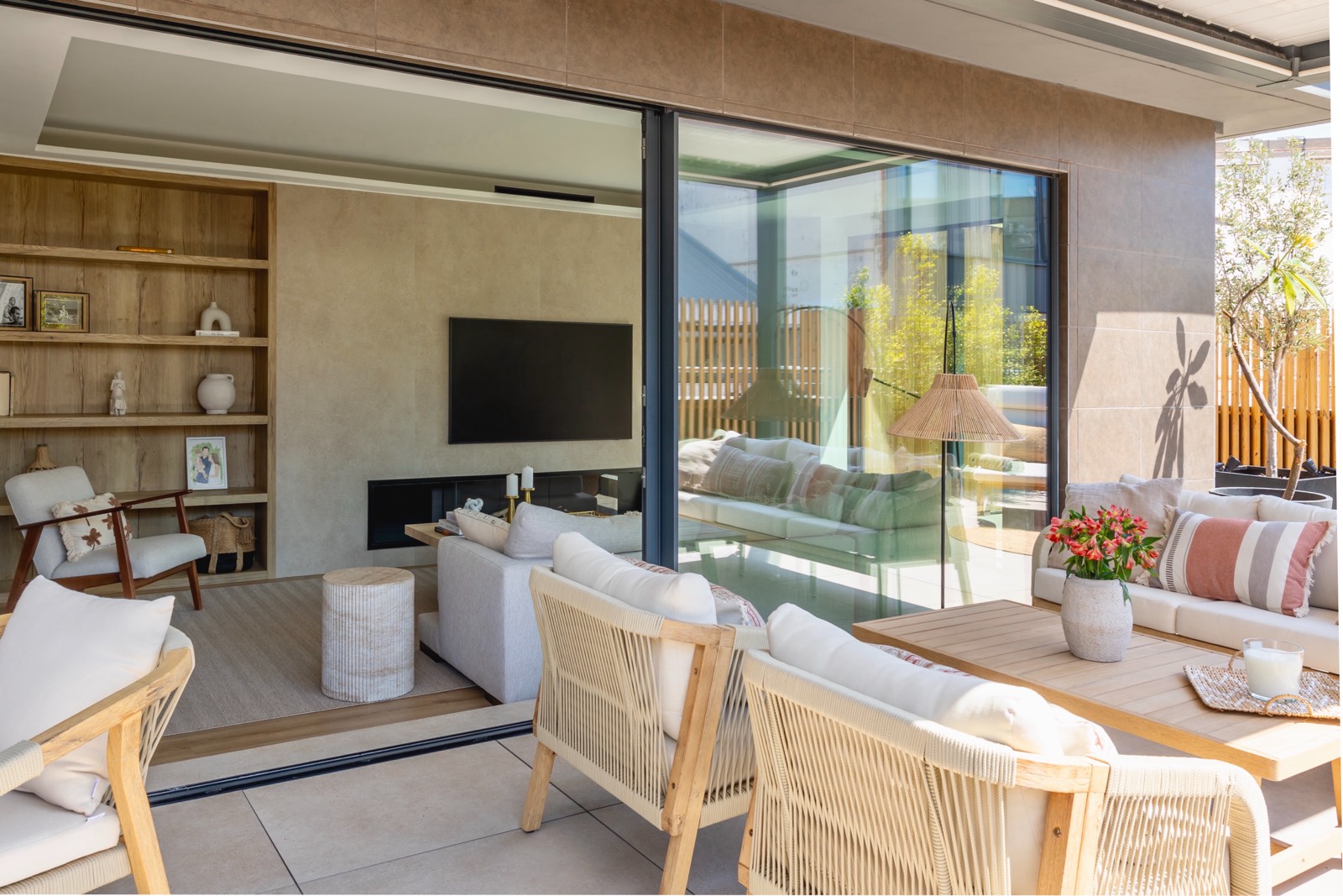 Salón moderno y luminoso con puertas correderas de cristal que se abren a una terraza acogedora. El interior cuenta con estanterías de madera, sofás blancos y una televisión. La terraza tiene muebles de exterior y plantas.