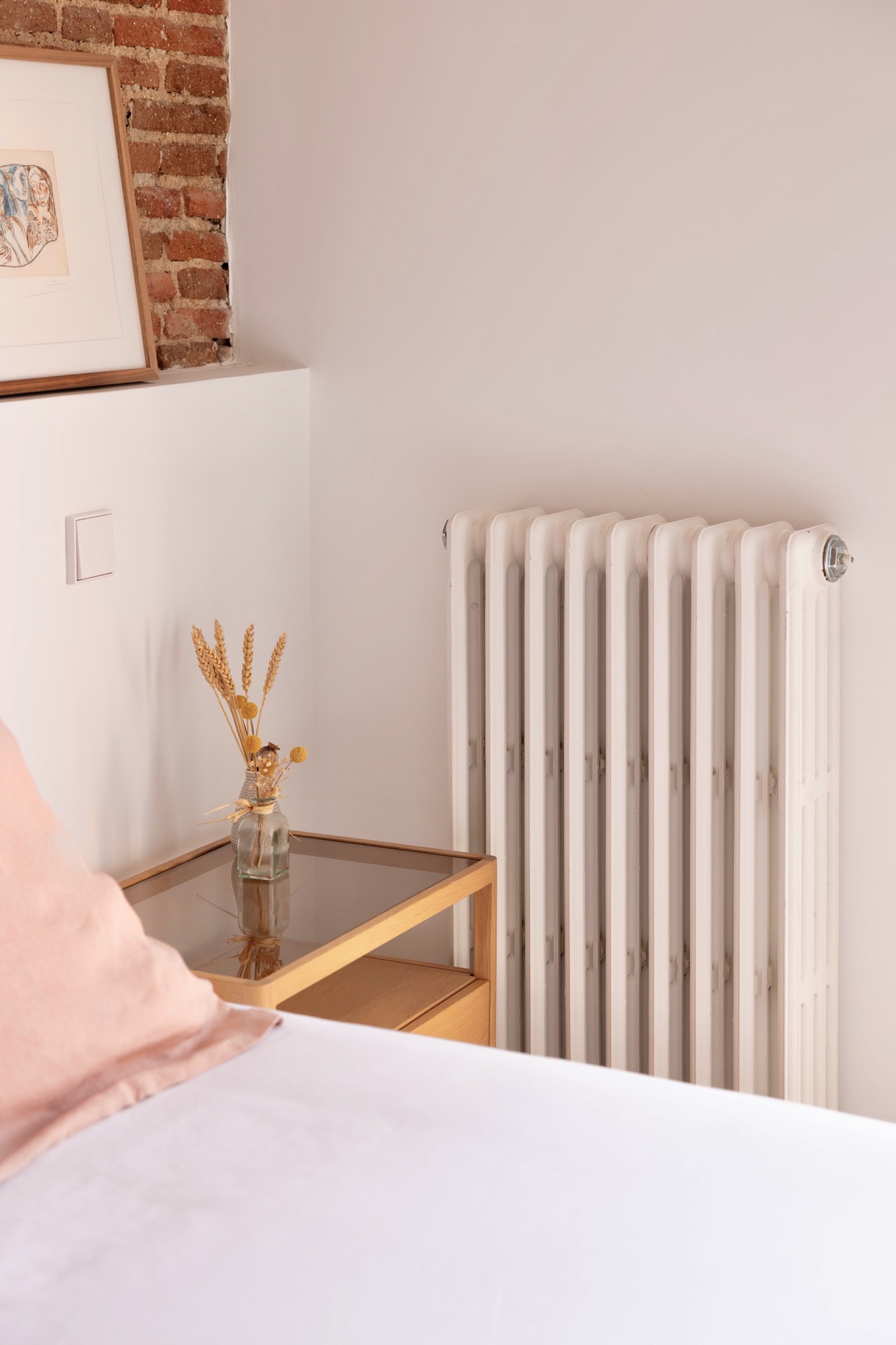 Interior de habitación con paredes blancas, radiador clásico y jarrón de cristal con espigas de trigo sobre mesita de noche de madera