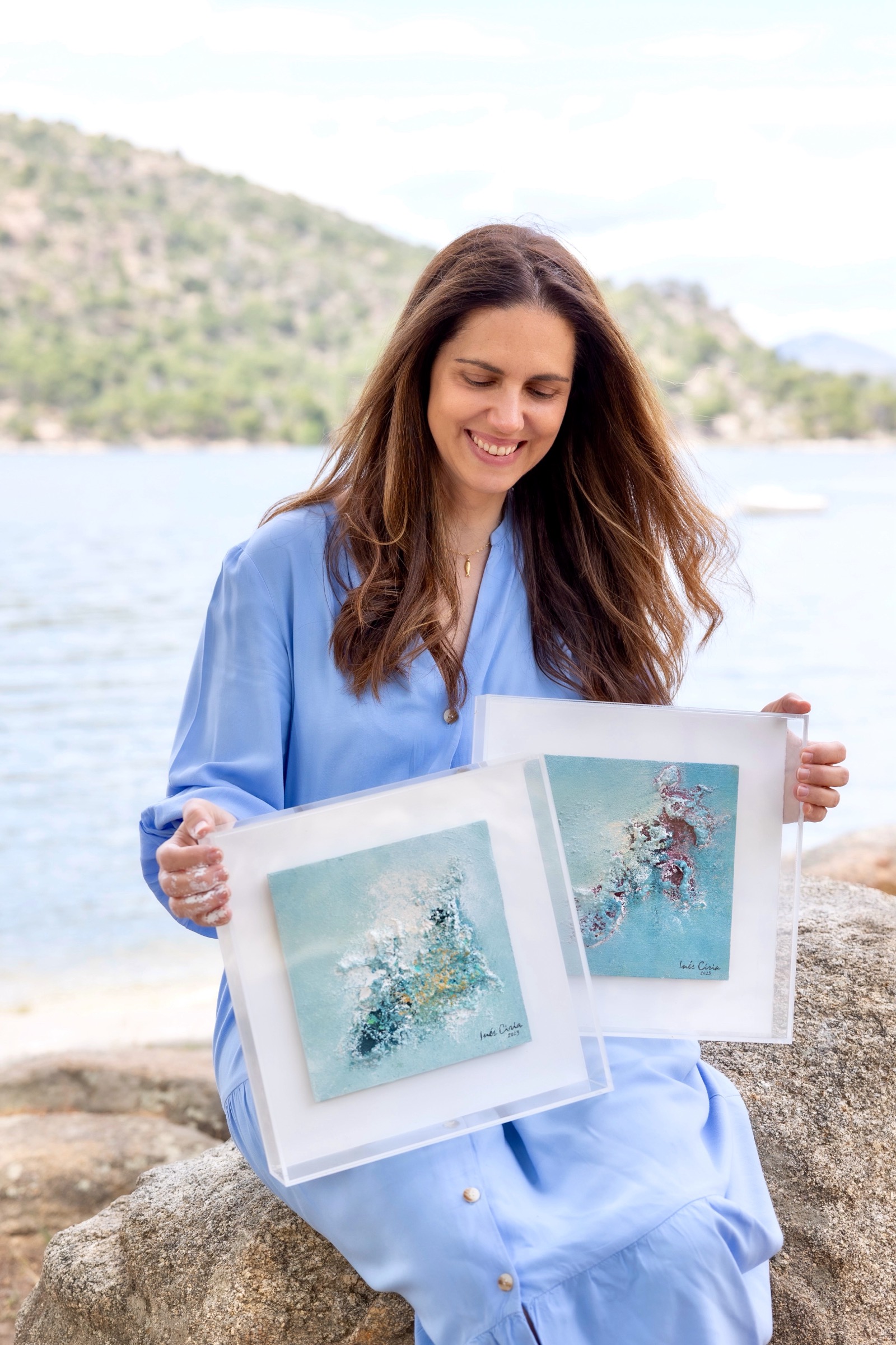 Mujer sonriente en vestido azul sentada en una roca junto al mar, sosteniendo dos pinturas abstractas en tonos azules y blancos. Montañas y agua en el fondo.