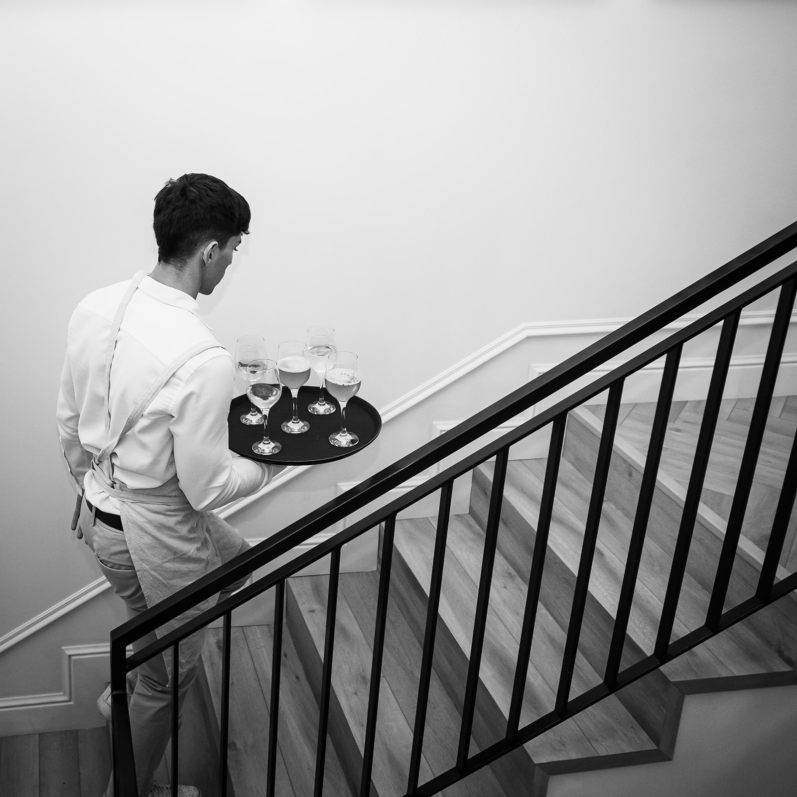 Imagen en blanco y negro de un camarero subiendo unas escaleras mientras lleva una bandeja redonda con seis copas de vino. El camarero viste una camisa blanca y un delantal, y está visto de espaldas.