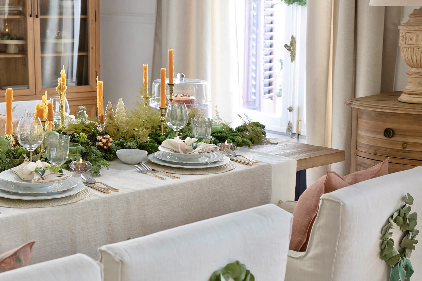 Mesa elegante decorada para Navidad con mantel beige, velas naranjas, elementos naturales como ramas de pino y eucalipto, vajilla blanca y copas de cristal. Sillas tapizadas en blanco con cojines rosa pálido.