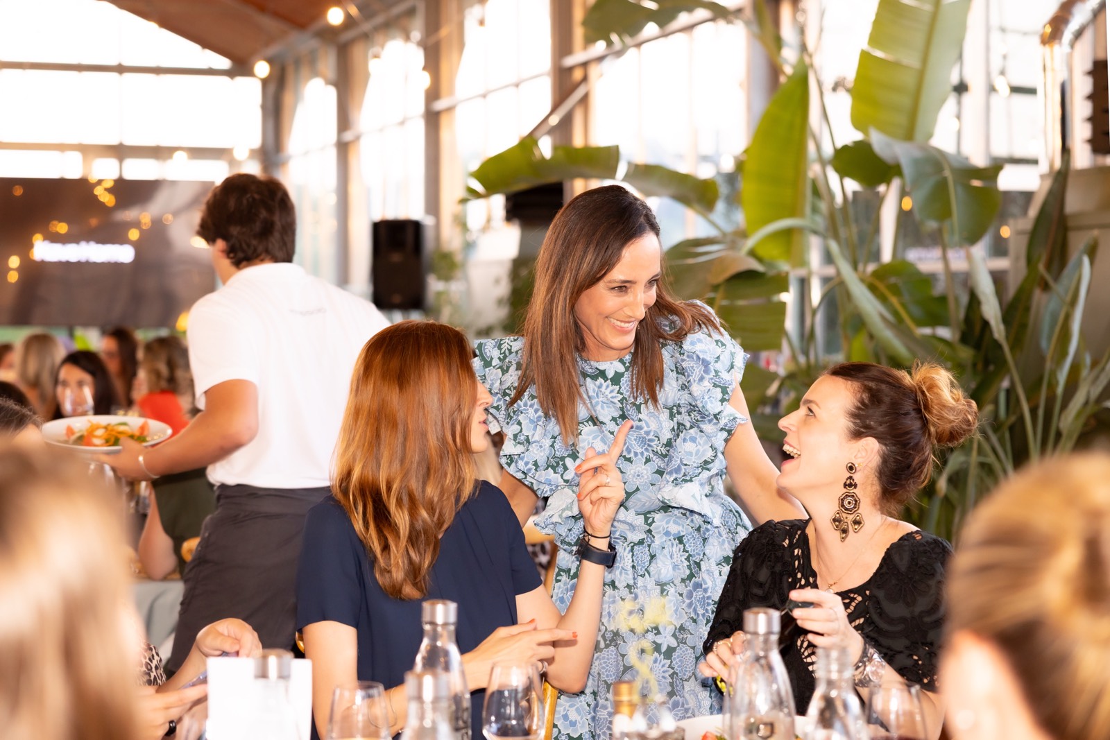 Grupo de mujeres sonrientes conversando animadamente en un restaurante con decoración de plantas y luces cálidas