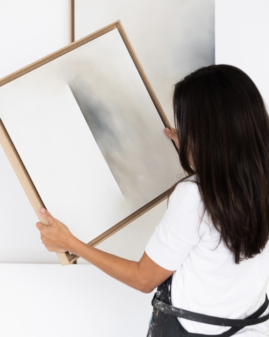 Mujer de espaldas sosteniendo un cuadro grande con marco de madera. Lleva una camiseta blanca y está en un espacio interior luminoso.