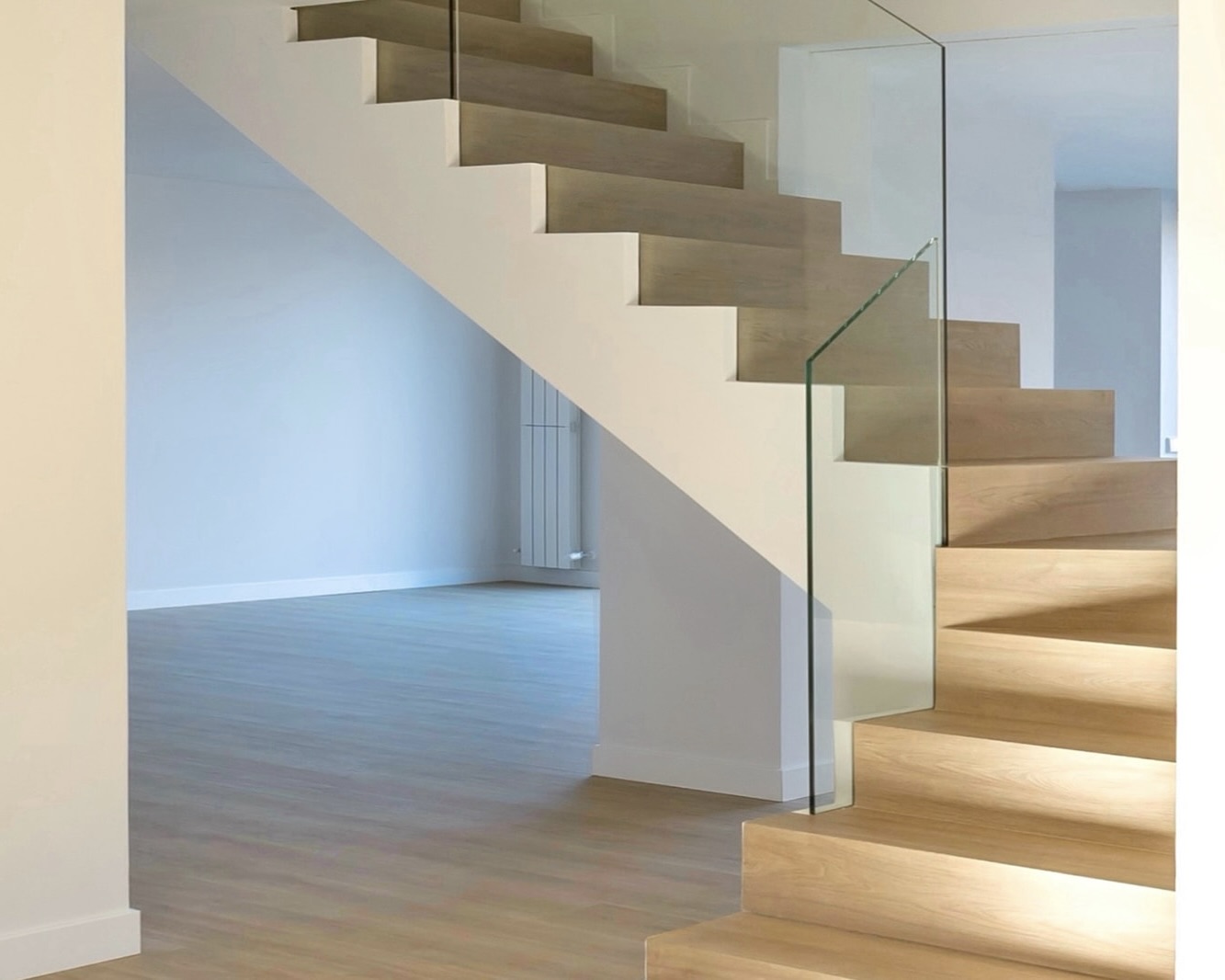 Escalera moderna de madera con barandilla de vidrio en un interior minimalista. Los escalones de madera clara contrastan con las paredes blancas y el suelo gris claro, creando un diseño elegante y luminoso.