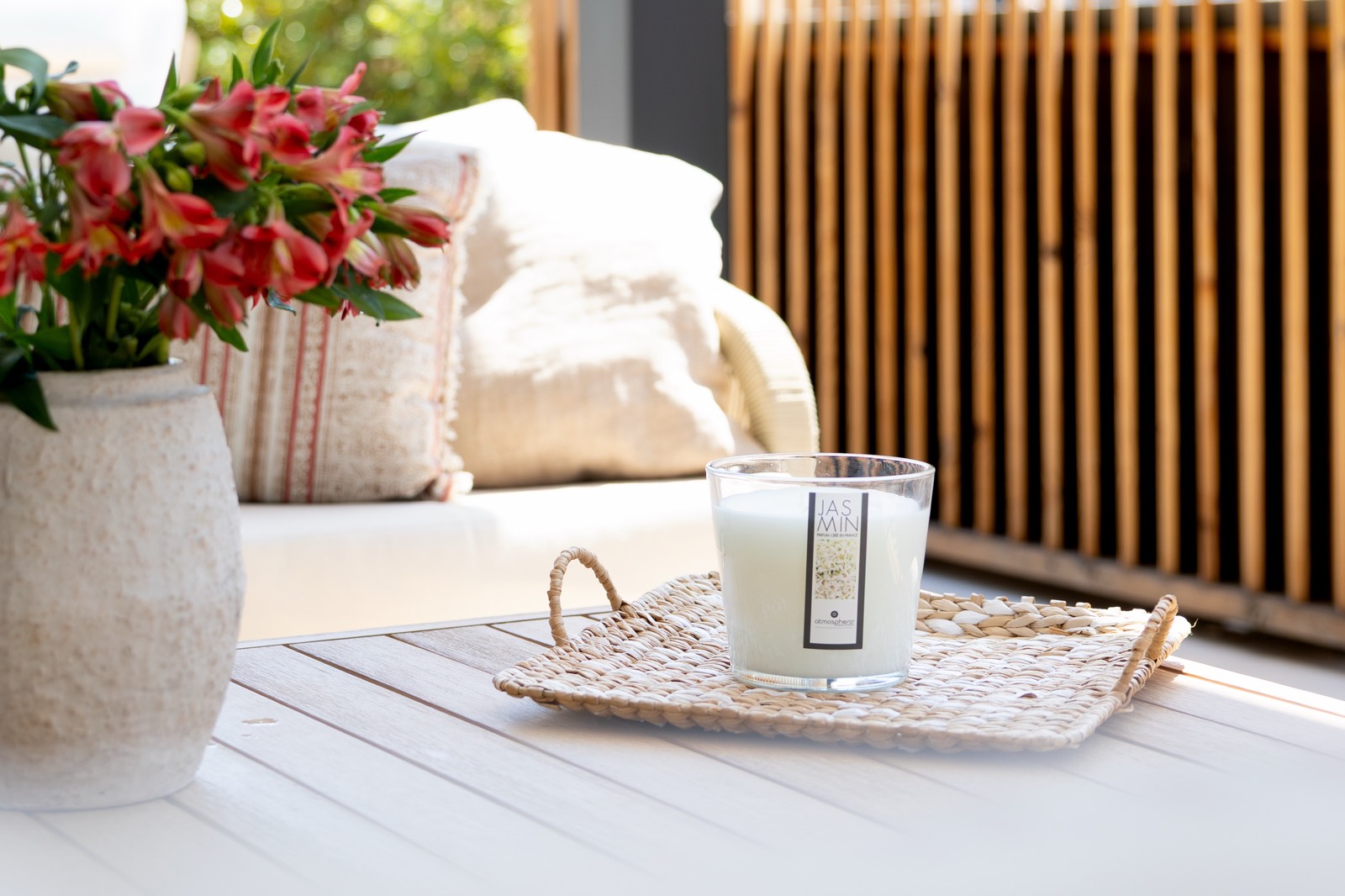Una terraza acogedora con una mesa blanca, un jarrón con flores rojas, una vela aromática sobre una bandeja de mimbre y cojines en un sofá al fondo. Se ve una pared de listones de madera.