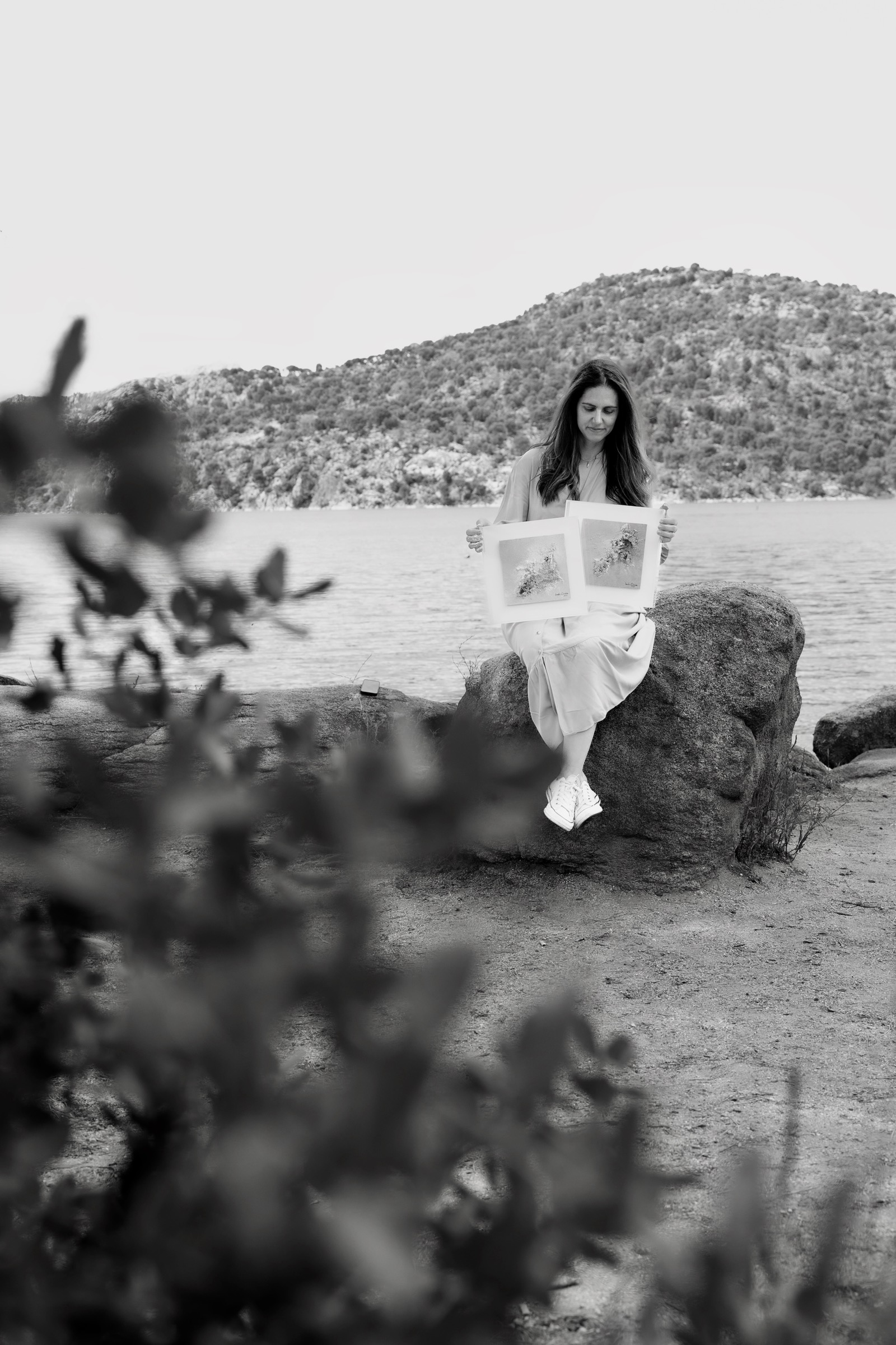 Imagen en blanco y negro de una persona sentada en una roca junto a un lago, leyendo un libro. En el fondo se ve una colina boscosa y en primer plano hay vegetación borrosa.