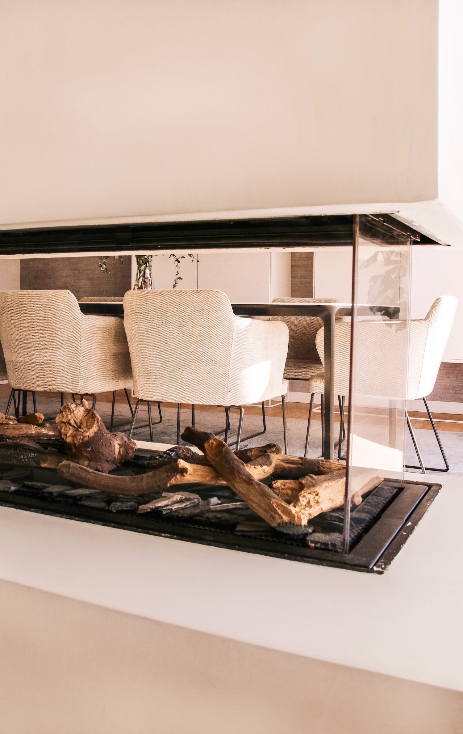 Comedor moderno con sillas tapizadas en tela beige, mesa de vidrio y decoración con troncos de madera natural en la base. Diseño minimalista en tonos neutros con acentos naturales.