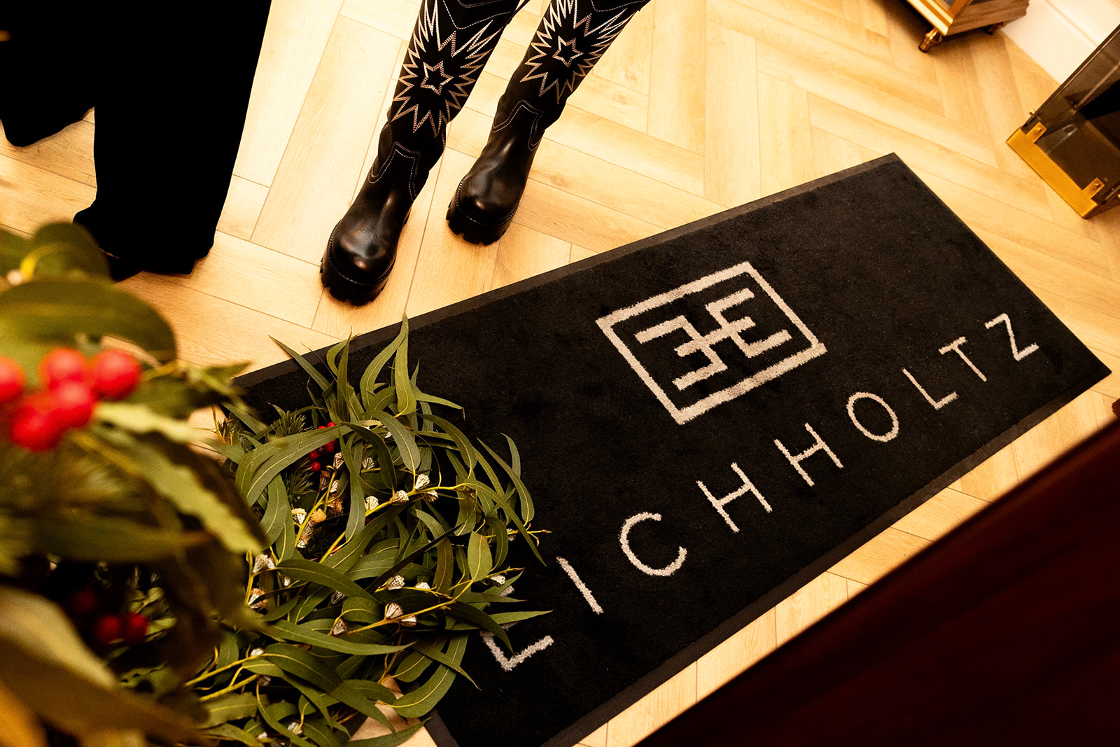 Alfombra negra con el logotipo y nombre 'LICHHOLTZ' en la entrada de una tienda. Se ven botas de cuero con diseño estrellado, plantas decorativas y suelo de madera.