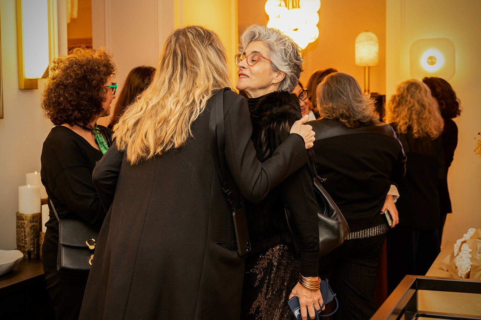 Grupo de mujeres elegantemente vestidas de negro en un evento social, conversando en un ambiente cálido e iluminado. La escena muestra interacciones cercanas y una atmósfera sofisticada.