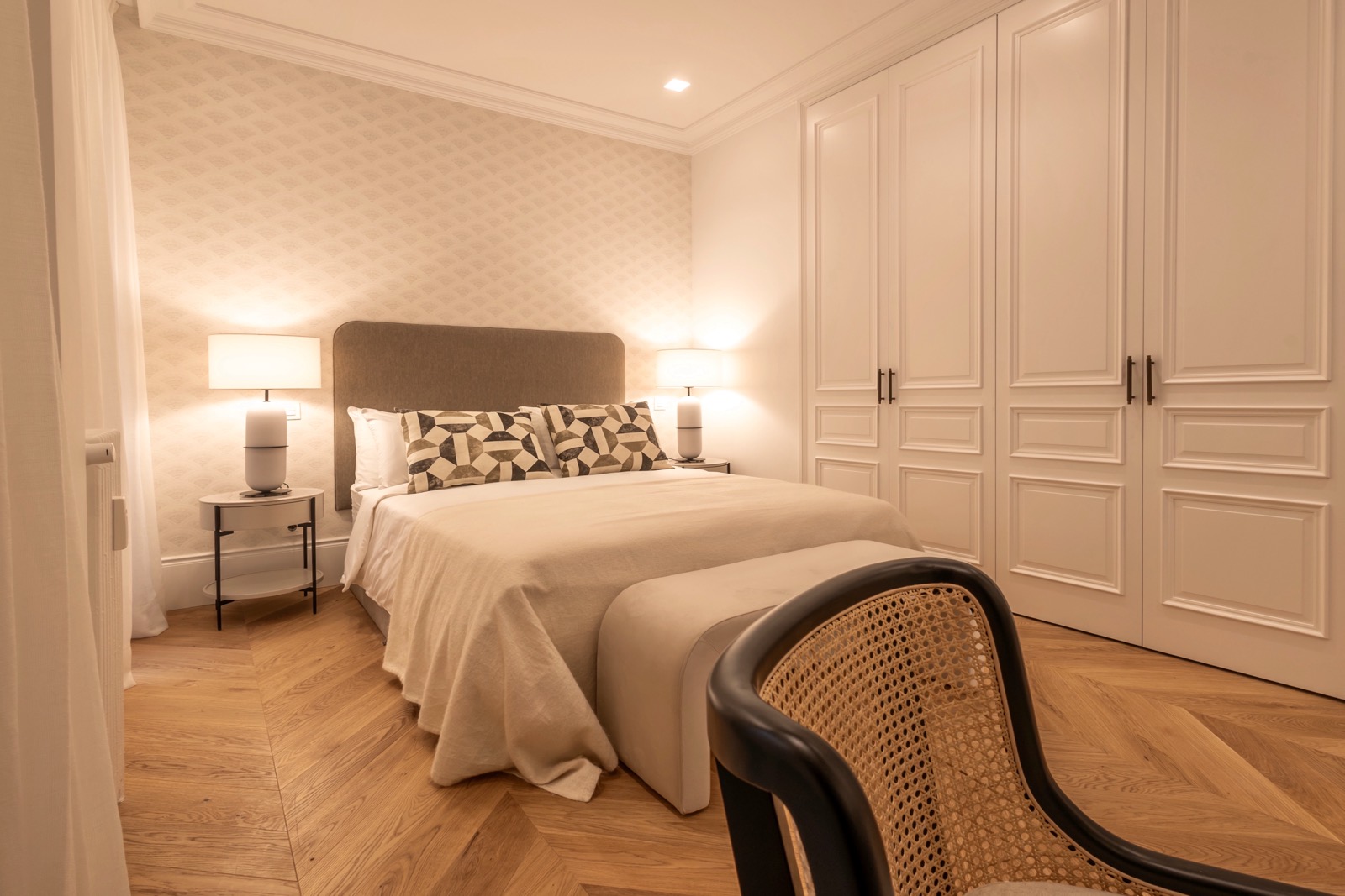 Dormitorio elegante con cama doble, armarios empotrados blancos, suelo de parquet, silla de rejilla y lámparas de mesa. Decoración en tonos neutros con detalles clásicos y modernos.