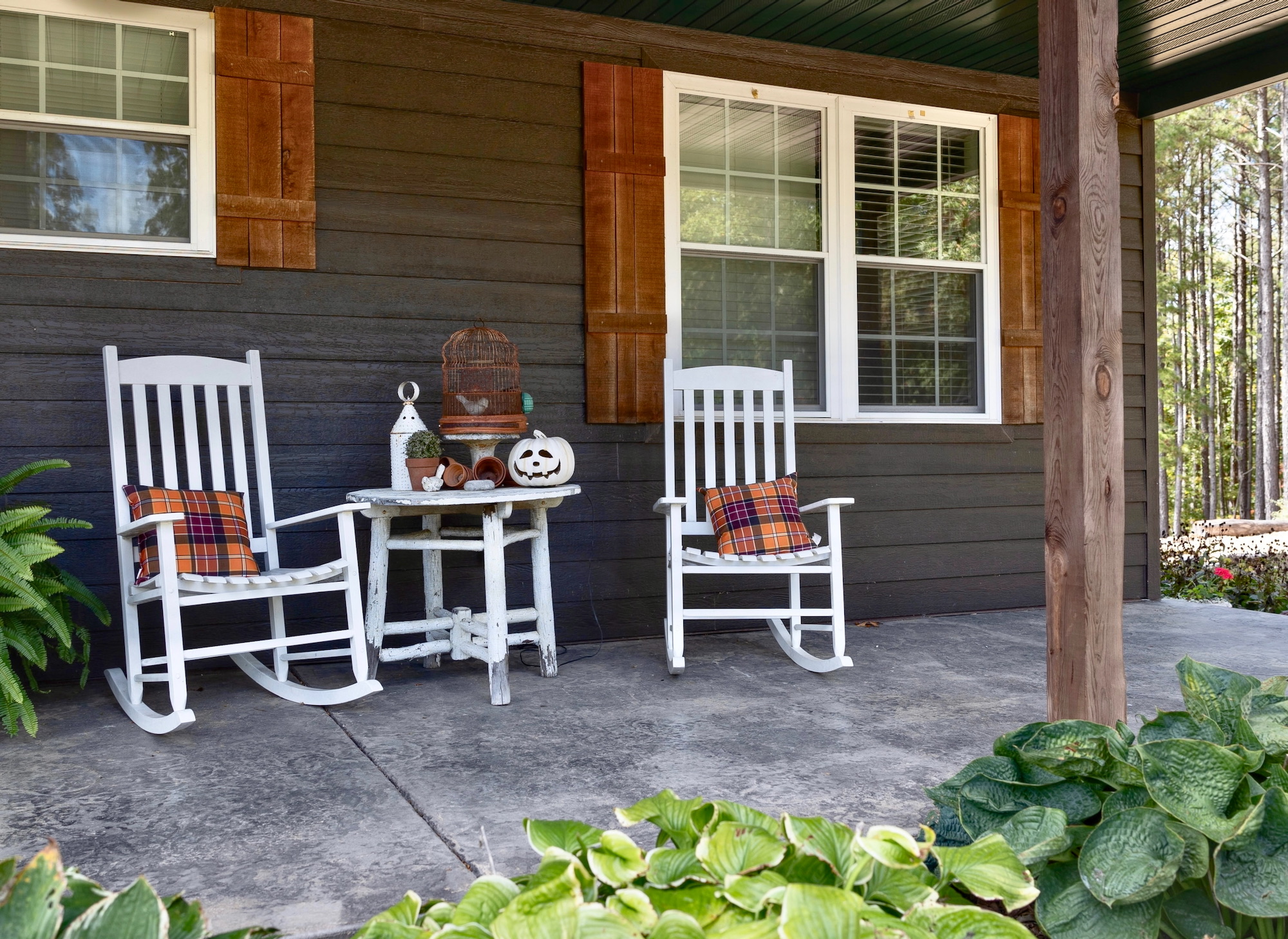 Porche acogedor de casa rústica con dos mecedoras blancas y una mesa pequeña, decorado con cojines a cuadros naranja, una calabaza decorativa y plantas. La fachada es de madera oscura con contraventanas marrones.