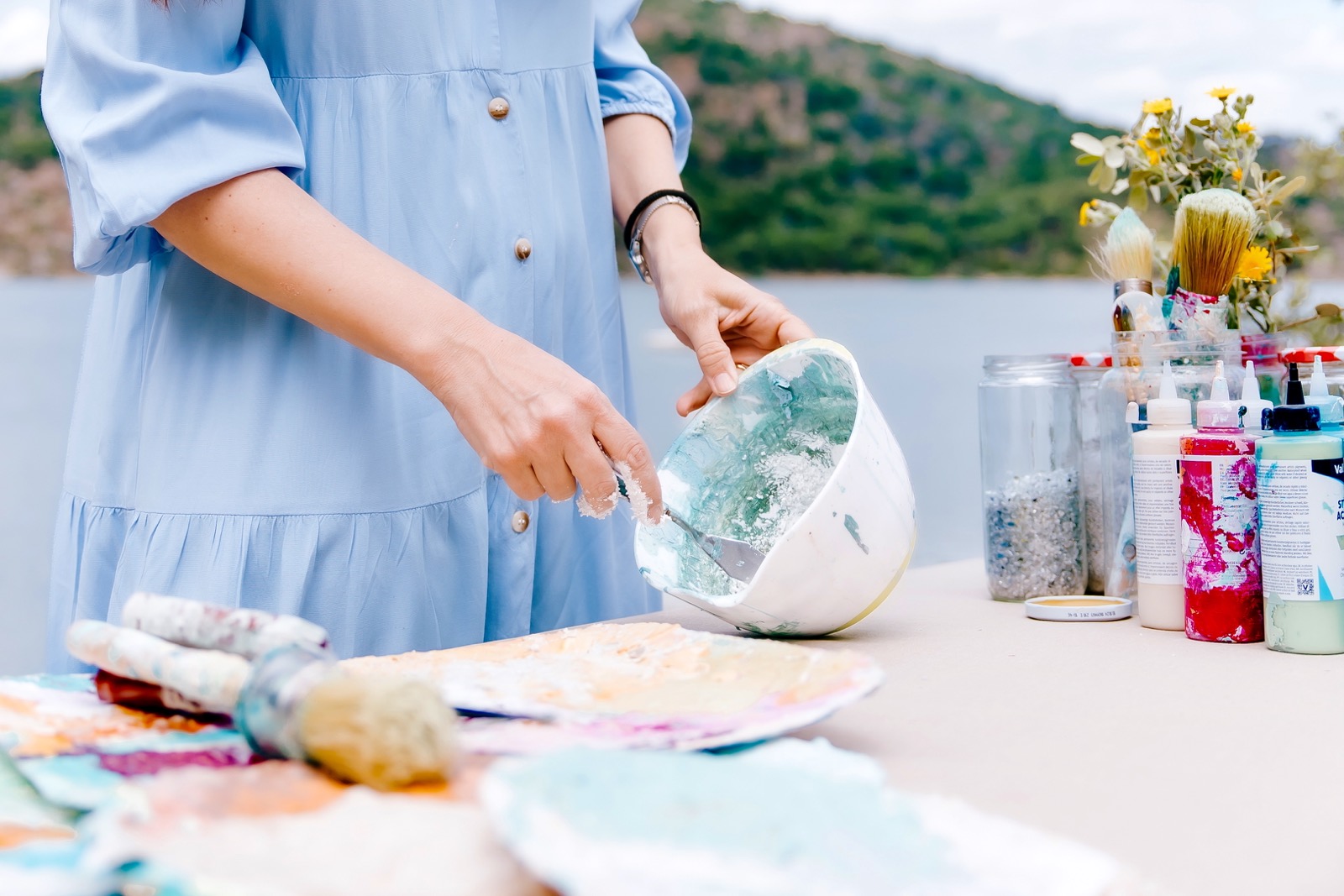 Una persona con vestido azul claro está pintando al aire libre. Se ve un tazón con pintura y varios materiales de arte sobre una mesa, con un paisaje montañoso y un lago de fondo.