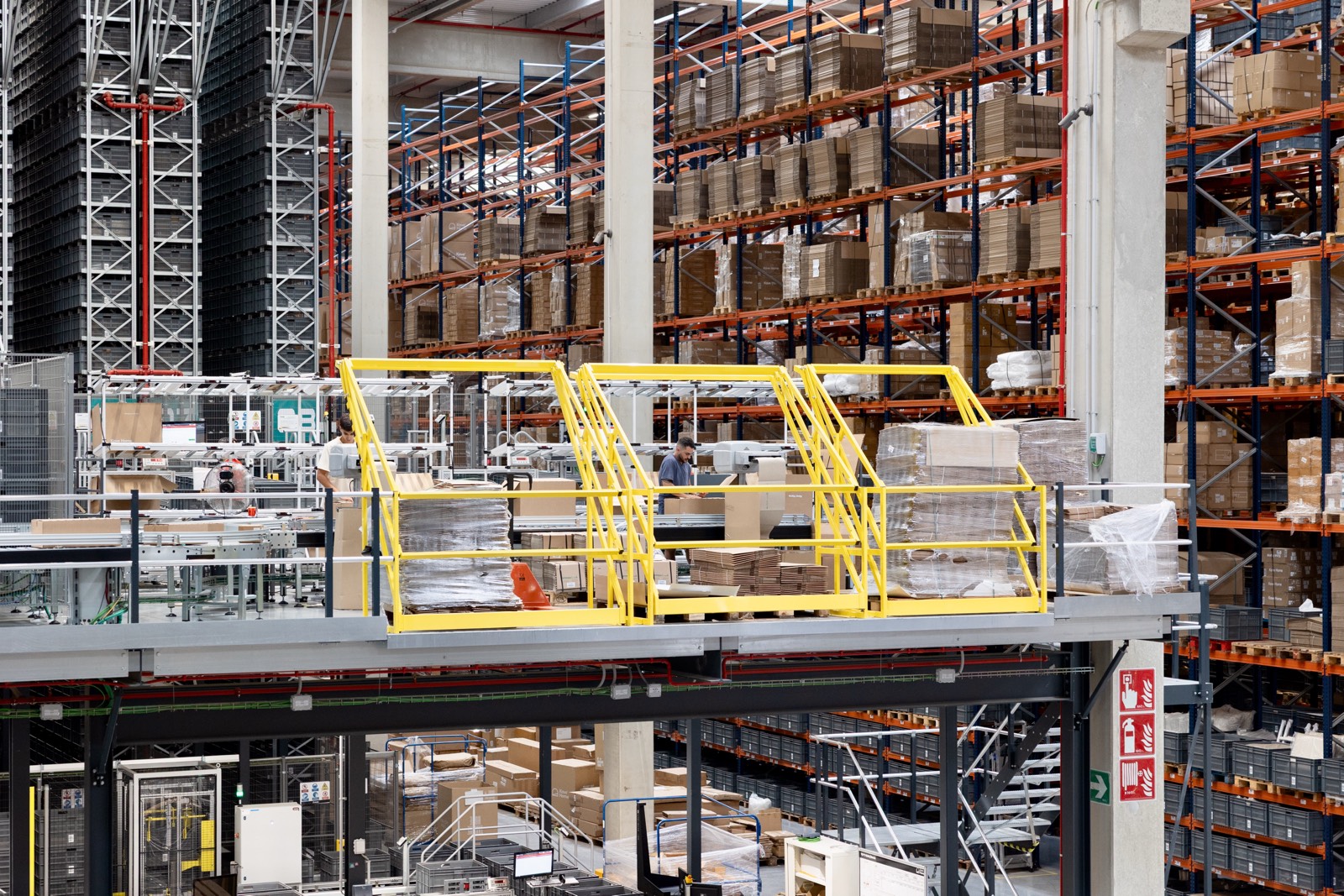 Interior de un gran almacén logístico con estanterías de múltiples niveles, plataformas amarillas y numerosas cajas y paquetes almacenados