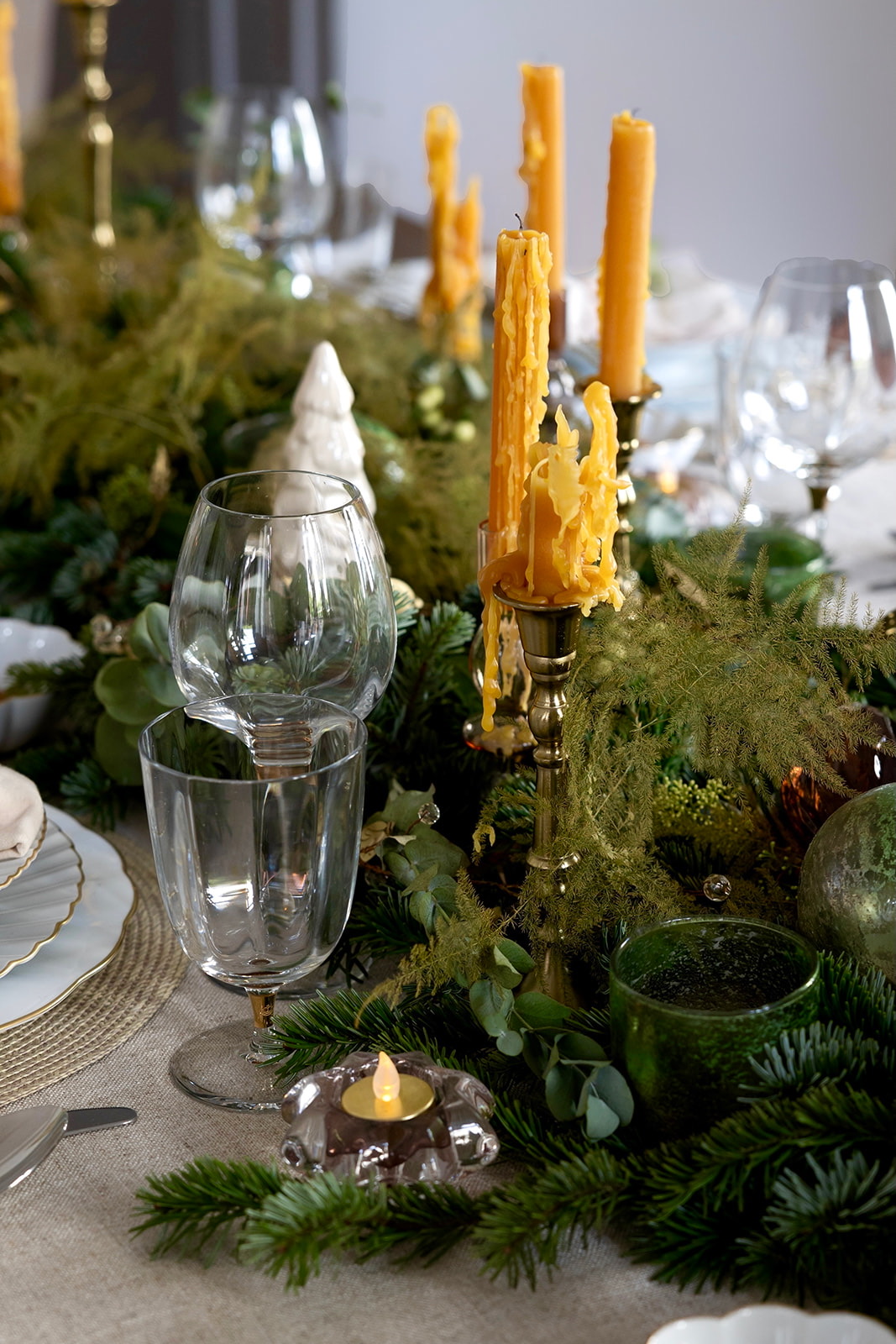 Decoración elegante de mesa navideña con velas amarillas derretidas en candelabros dorados, copas de cristal y guirnalda de ramas de pino y eucalipto. Centro de mesa festivo con adornos verdes y velas.