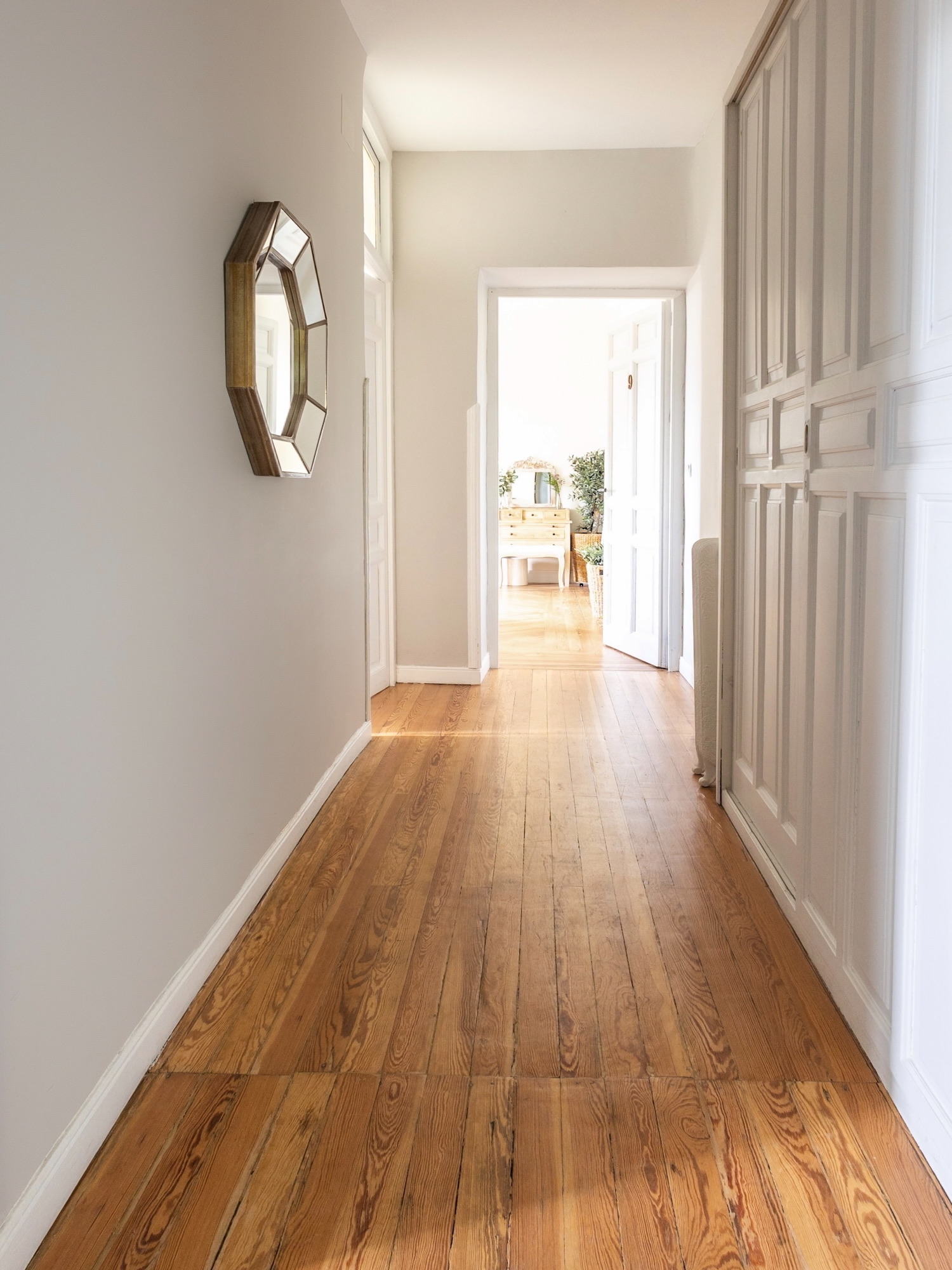 Pasillo largo y luminoso con paredes blancas, piso de madera clara pulida, espejo octagonal decorativo en la pared y armarios empotrados blancos. Al fondo se vislumbra una habitación iluminada por luz natural.