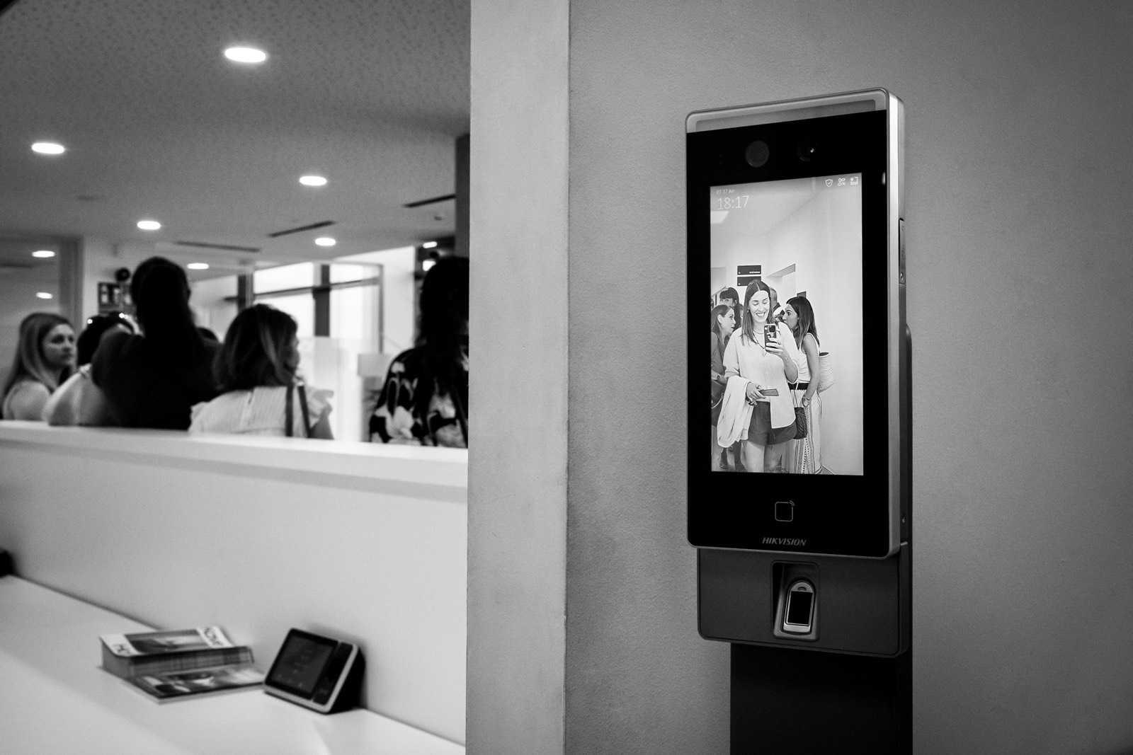 Pantalla de dispositivo Hikvision mostrando un grupo de personas reflejadas, con un área de recepción o entrada al fondo