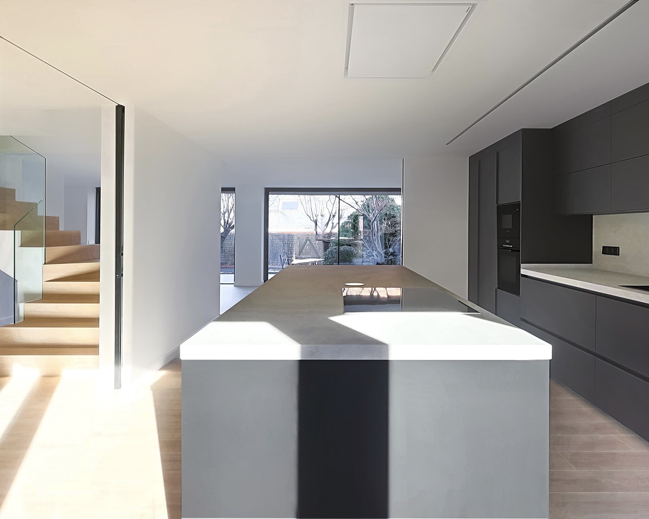 Cocina moderna y minimalista con isla central blanca, armarios grises, ventanal grande y escaleras de madera visibles. El diseño es limpio y luminoso con un ambiente abierto y espacioso.