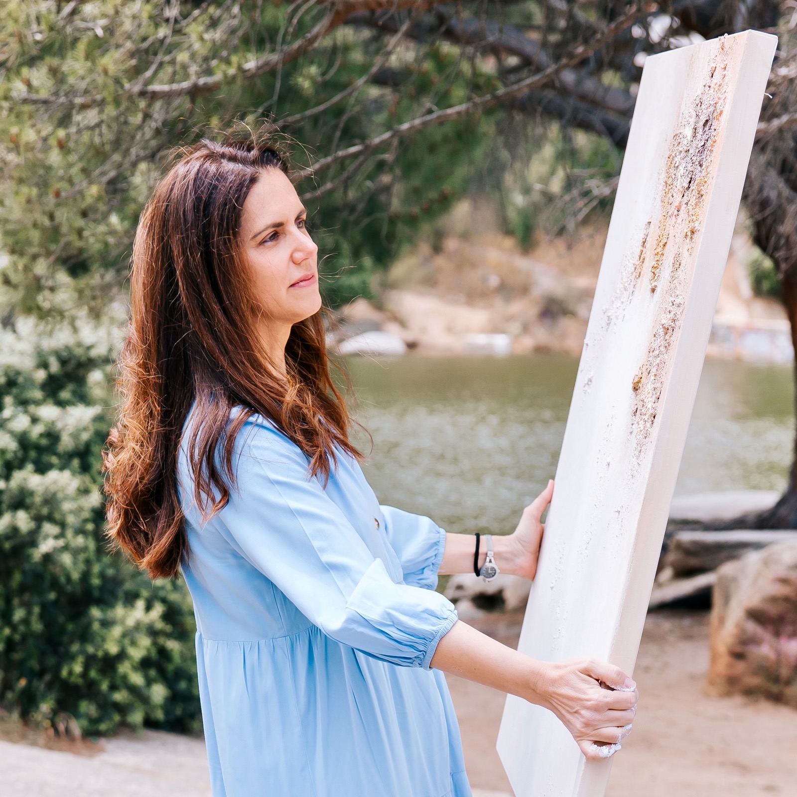 Mujer con vestido azul sosteniendo un lienzo en blanco frente a un lago, rodeada de naturaleza. Su expresión es concentrada mientras observa el paisaje, como si estuviera a punto de comenzar a pintar.