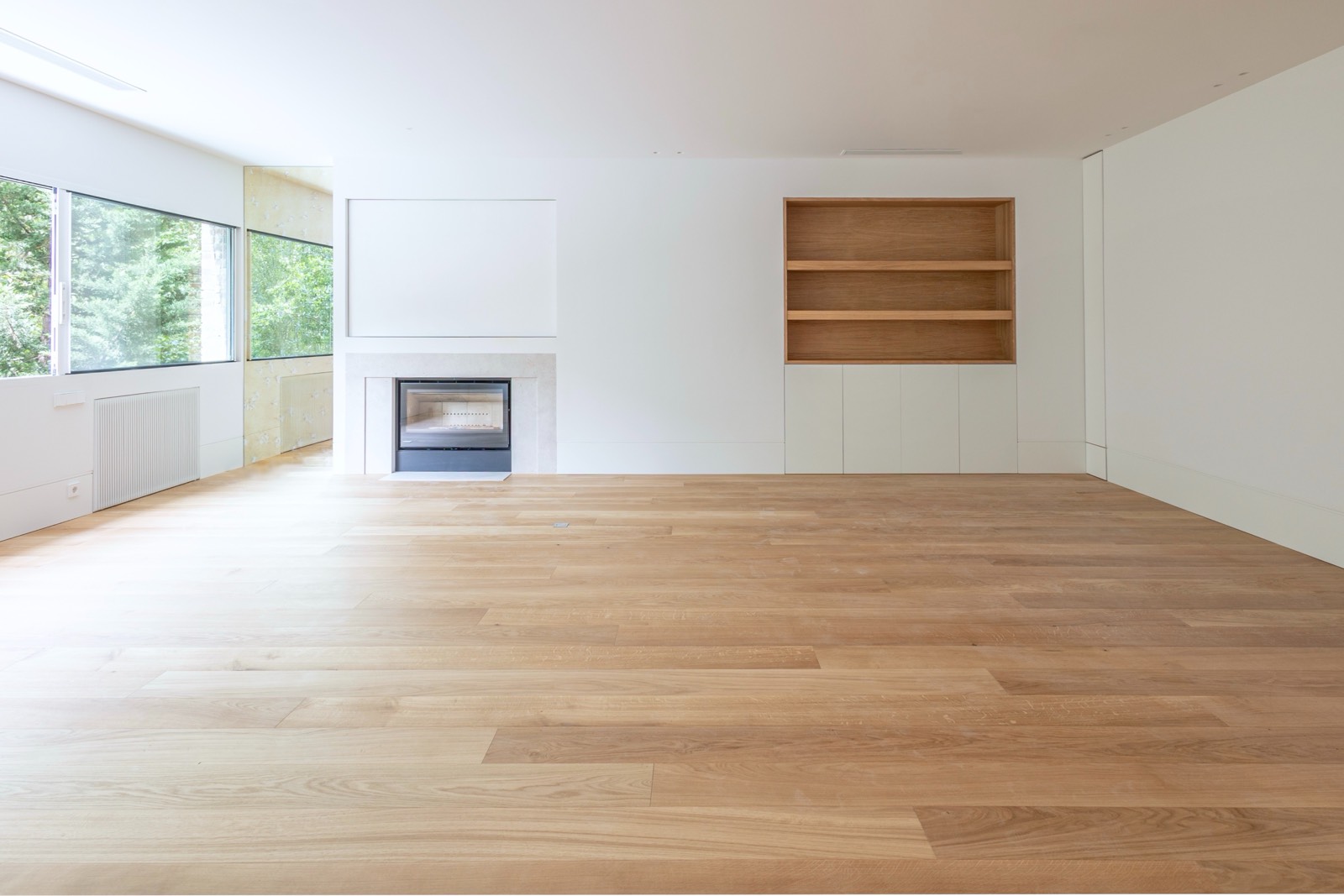 Interior de habitación vacía con suelo de madera clara, paredes blancas, ventanas grandes y chimenea minimalista con estantería de madera