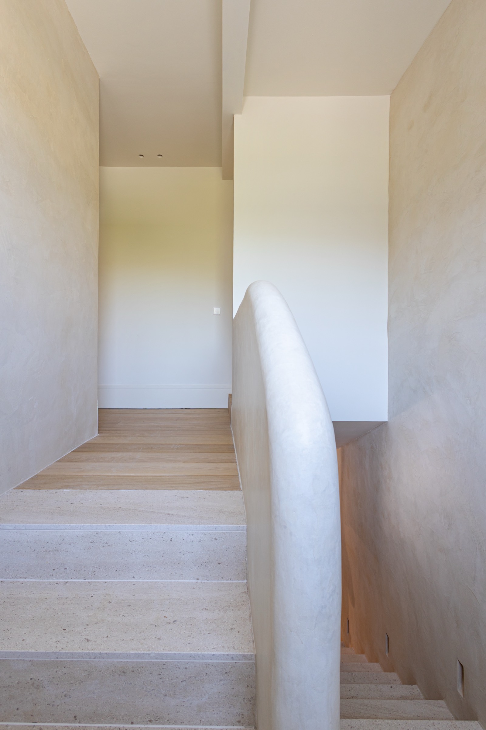 Tramo de escalera interior con paredes beige claras, peldaños de piedra y madera, y barandilla blanca curva de diseño moderno