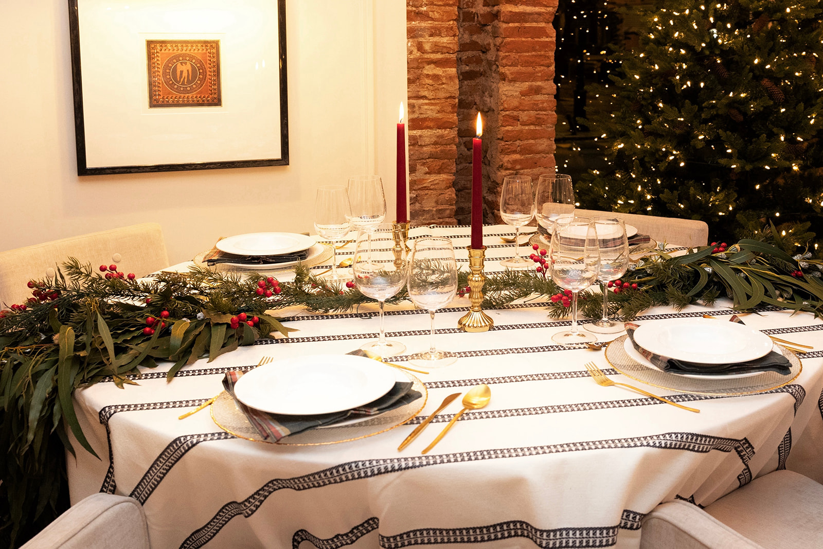 Mesa elegantemente decorada para una cena navideña con un mantel blanco, velas rojas, guirnaldas verdes con bayas rojas, copas de vino y platos blancos. Al fondo se ve parte de un árbol de Navidad iluminado.