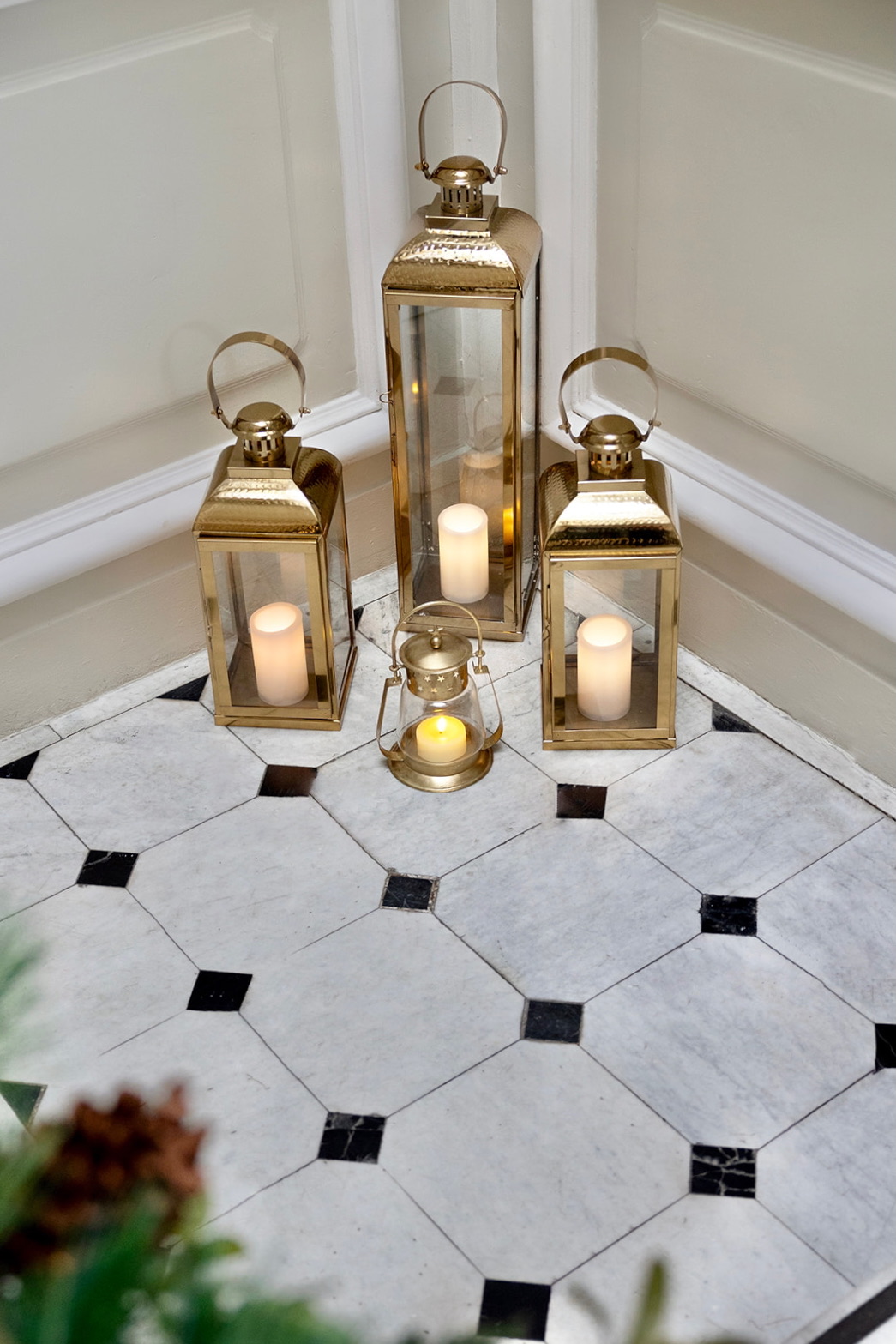 Conjunto de faroles dorados decorativos con velas LED sobre suelo de mármol blanco y negro en patrón octagonal. Los faroles son de diferentes tamaños y están agrupados elegantemente en una esquina.