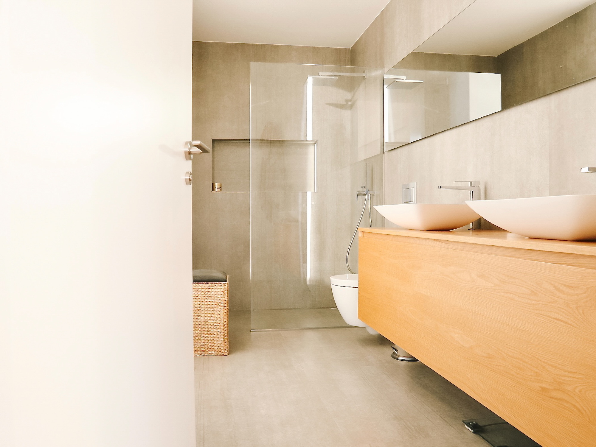 Baño moderno con mueble flotante de madera clara, lavabos blancos tipo bowl, ducha con mampara de cristal, paredes en tonos beige y espejo horizontal. Diseño minimalista con cesta de mimbre decorativa.