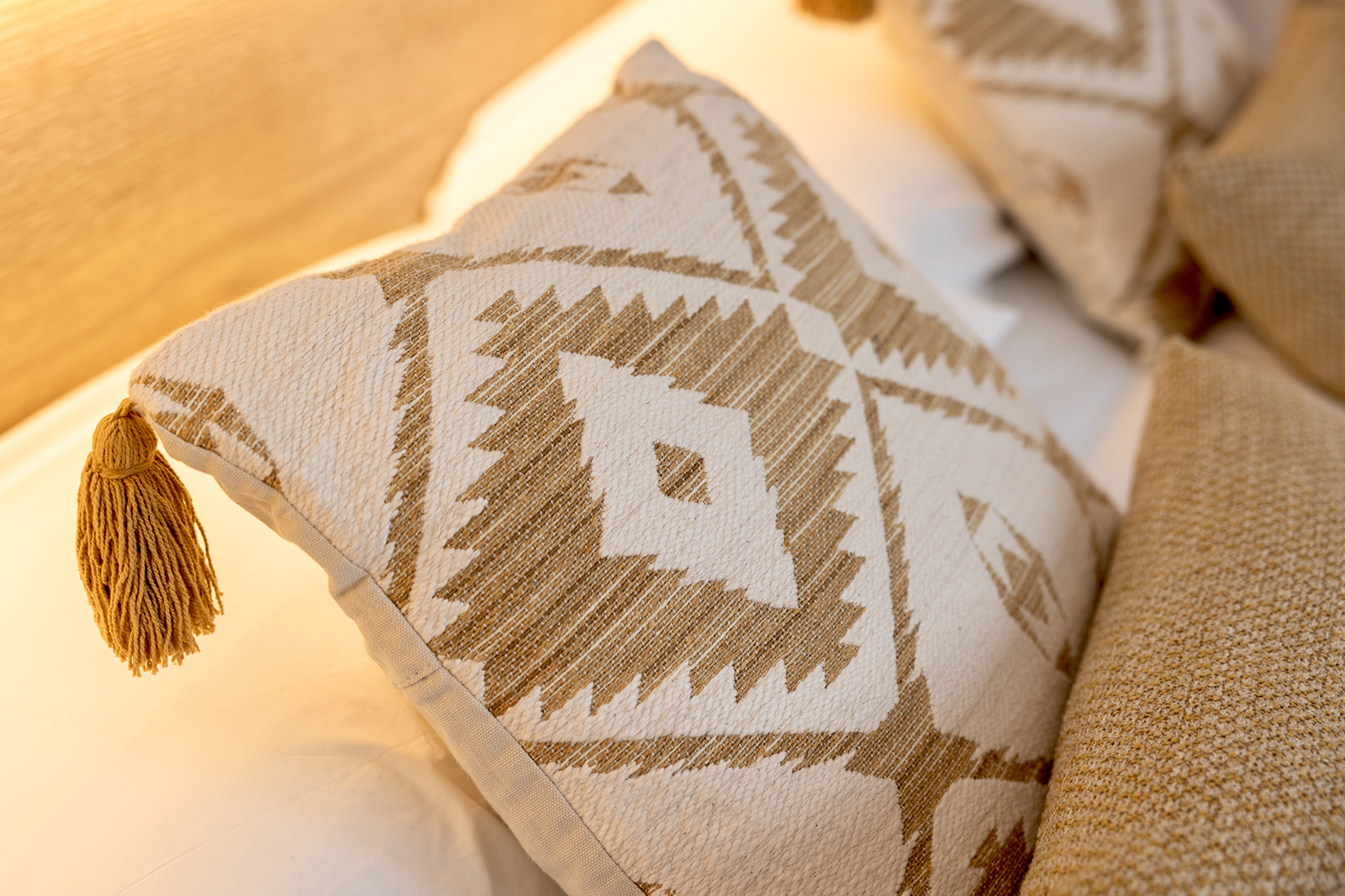 Cojín decorativo con diseño geométrico en tonos beige y marrón, estilo boho, con borla dorada en una esquina, sobre un sofá de color crema iluminado por luz cálida