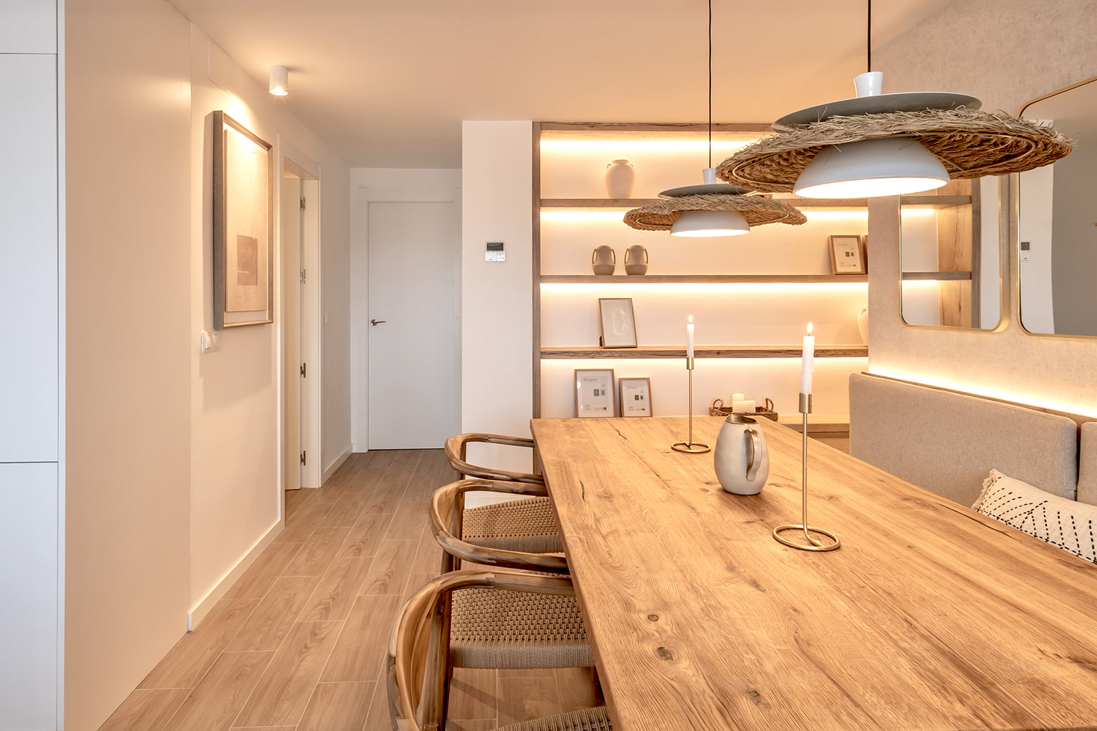 Comedor moderno de estilo escandinavo con mesa de madera, sillas de ratán, lámparas colgantes tejidas y estanterías iluminadas. Espacio luminoso y acogedor con tonos cálidos y elementos naturales.