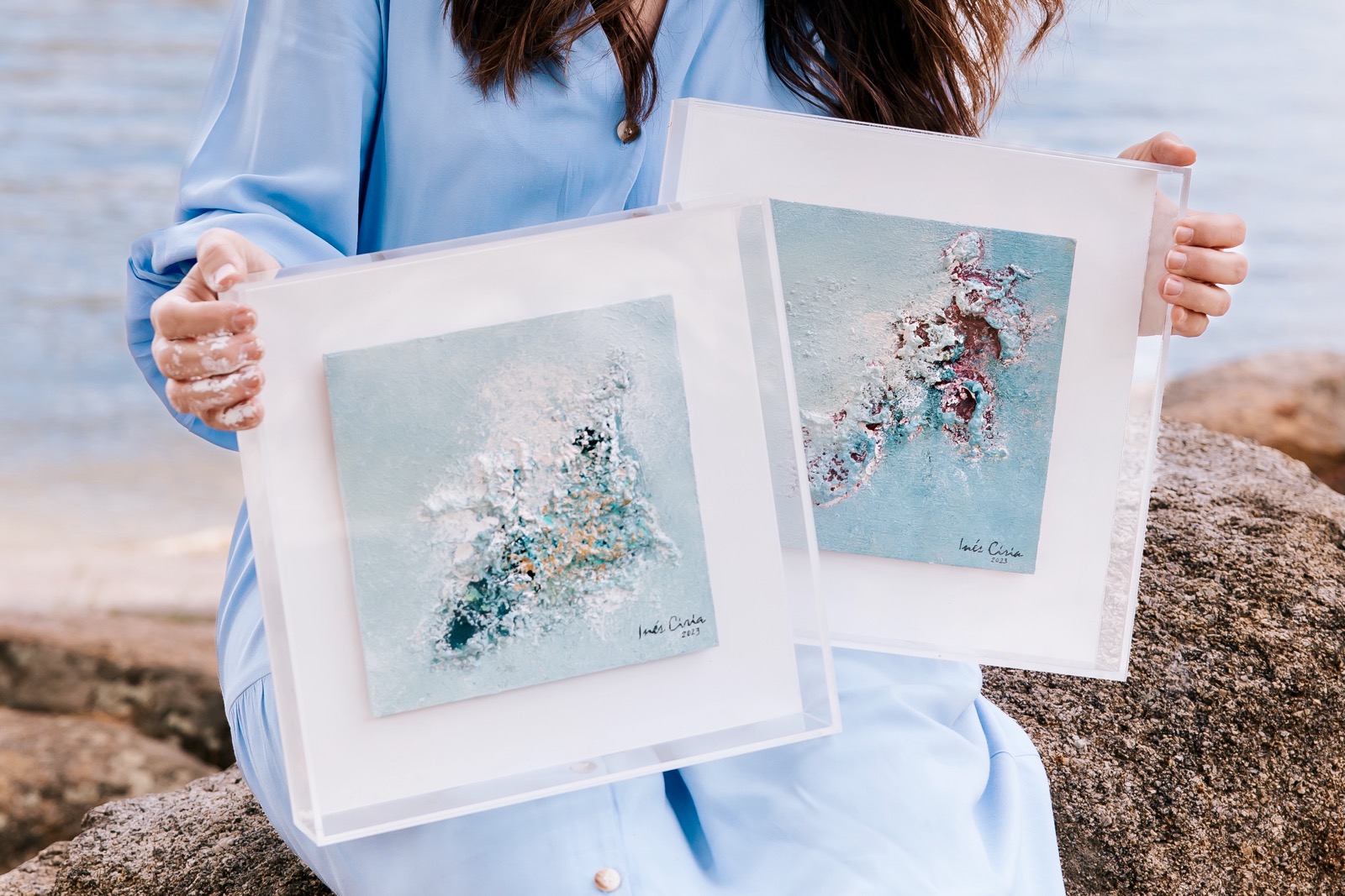 Persona sosteniendo dos cuadros de arte abstracto con temas marinos en tonos azules y blancos junto a una orilla rocosa