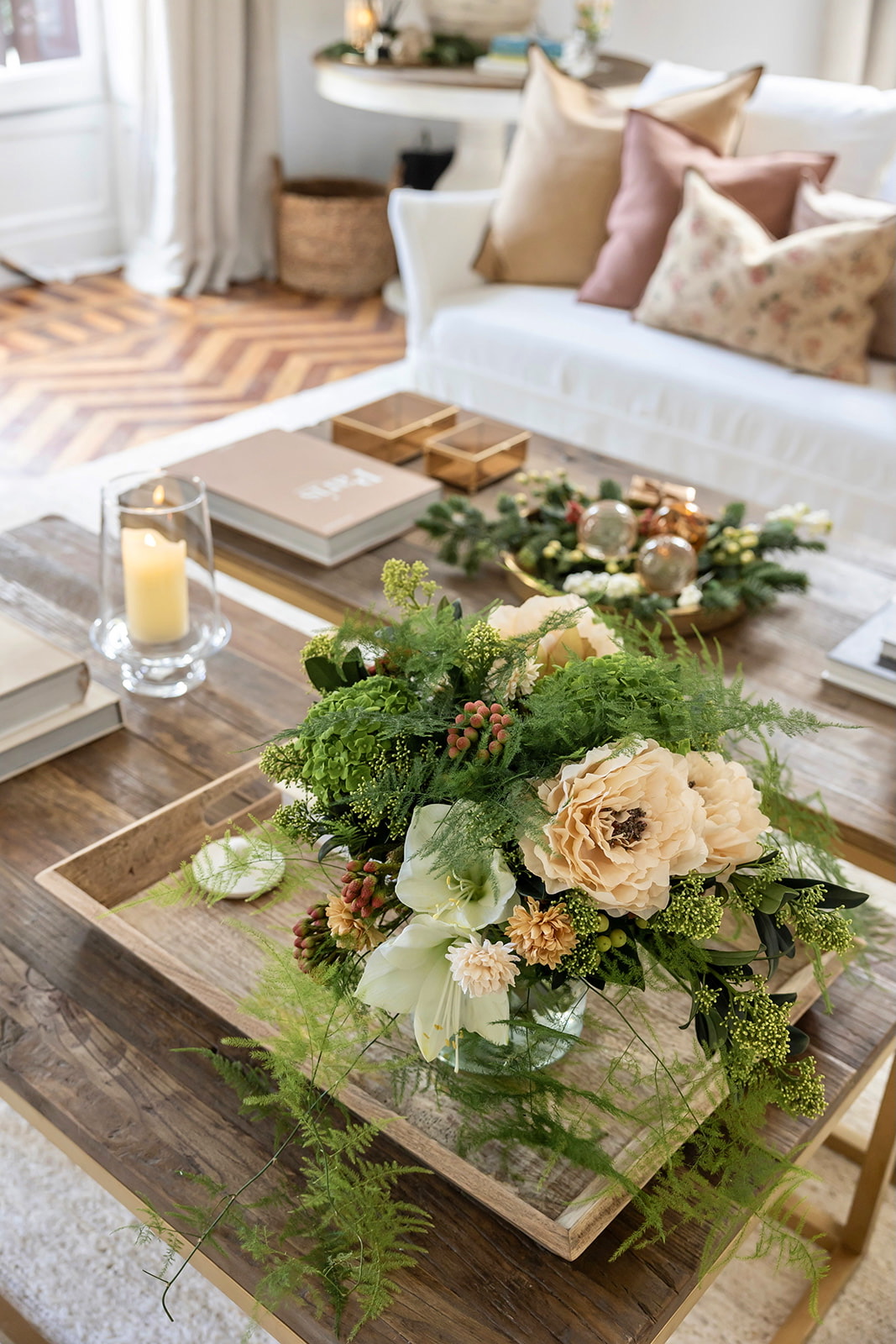 Elegante arreglo floral en tonos crema y verde sobre mesa de café de madera, acompañado de velas y libros decorativos. El ambiente muestra un sofá blanco con cojines en tonos nude y rosa, creando una atmósfera acogedora