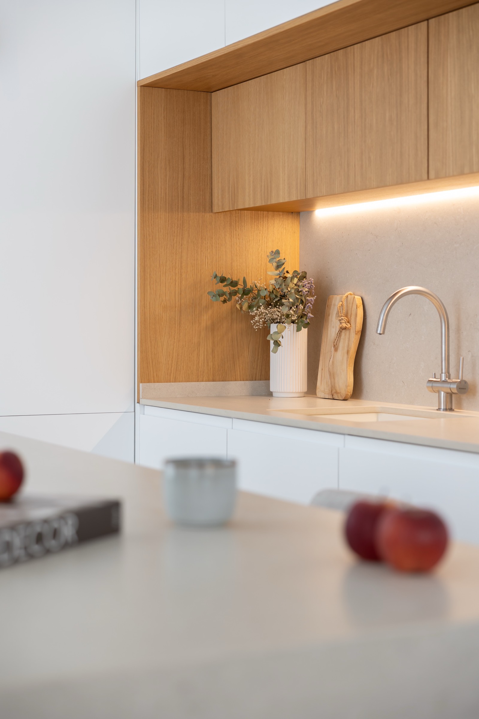 Cocina moderna con gabinetes de madera clara y blancos, encimera blanca, grifo de acero inoxidable, iluminación bajo muebles, y detalles decorativos como un jarrón con flores y una tabla de cortar