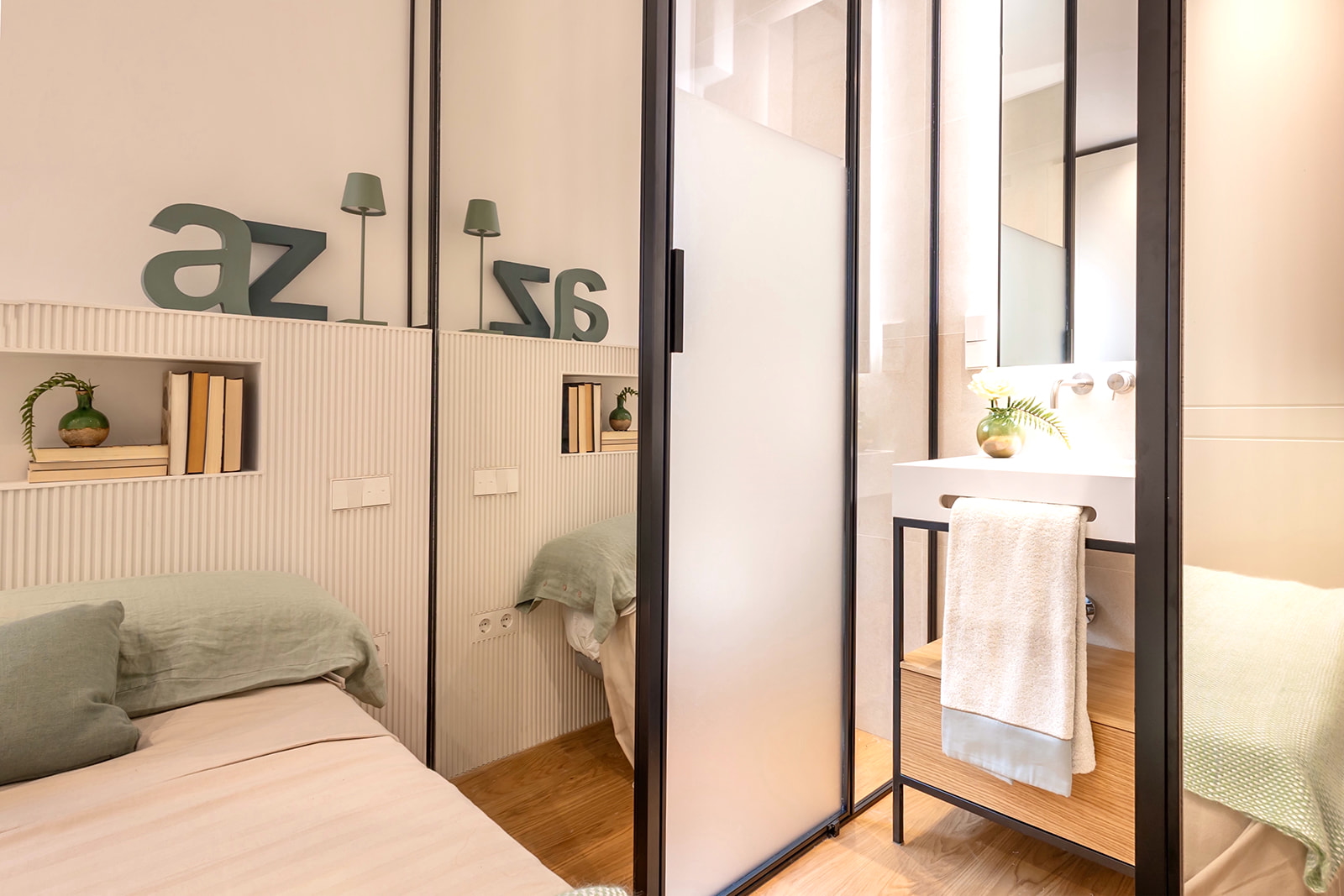 Dormitorio moderno con cabecero decorado con letras 'az', estantes empotrados y baño integrado separado por puerta corredera de cristal. Diseño minimalista en tonos neutros y verdes.
