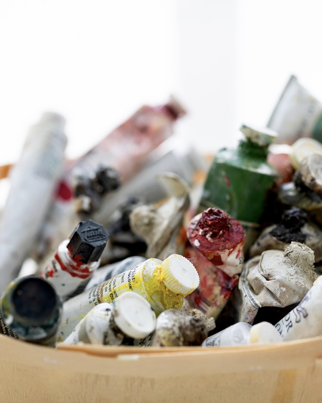 Primer plano de una colección de tubos de pintura usados y desgastados en diversos colores, apilados desordenadamente en un recipiente. Los tubos muestran signos de uso intenso, con pintura seca y manchas en sus superficies.