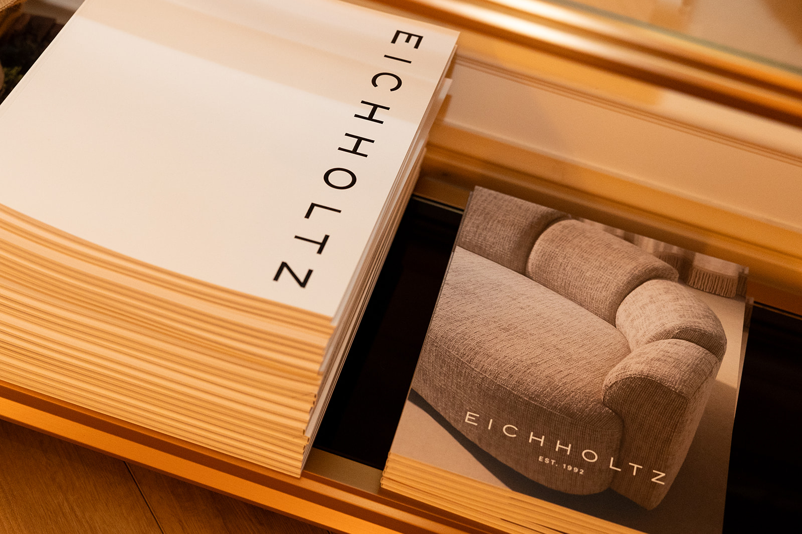 Catálogo de Eichholtz mostrando una pila de libros con el nombre de la marca y una imagen de un sofá elegante en tonos beige. Los libros están sobre una superficie de madera, sugiriendo un ambiente de diseño de interiores de lujo.
