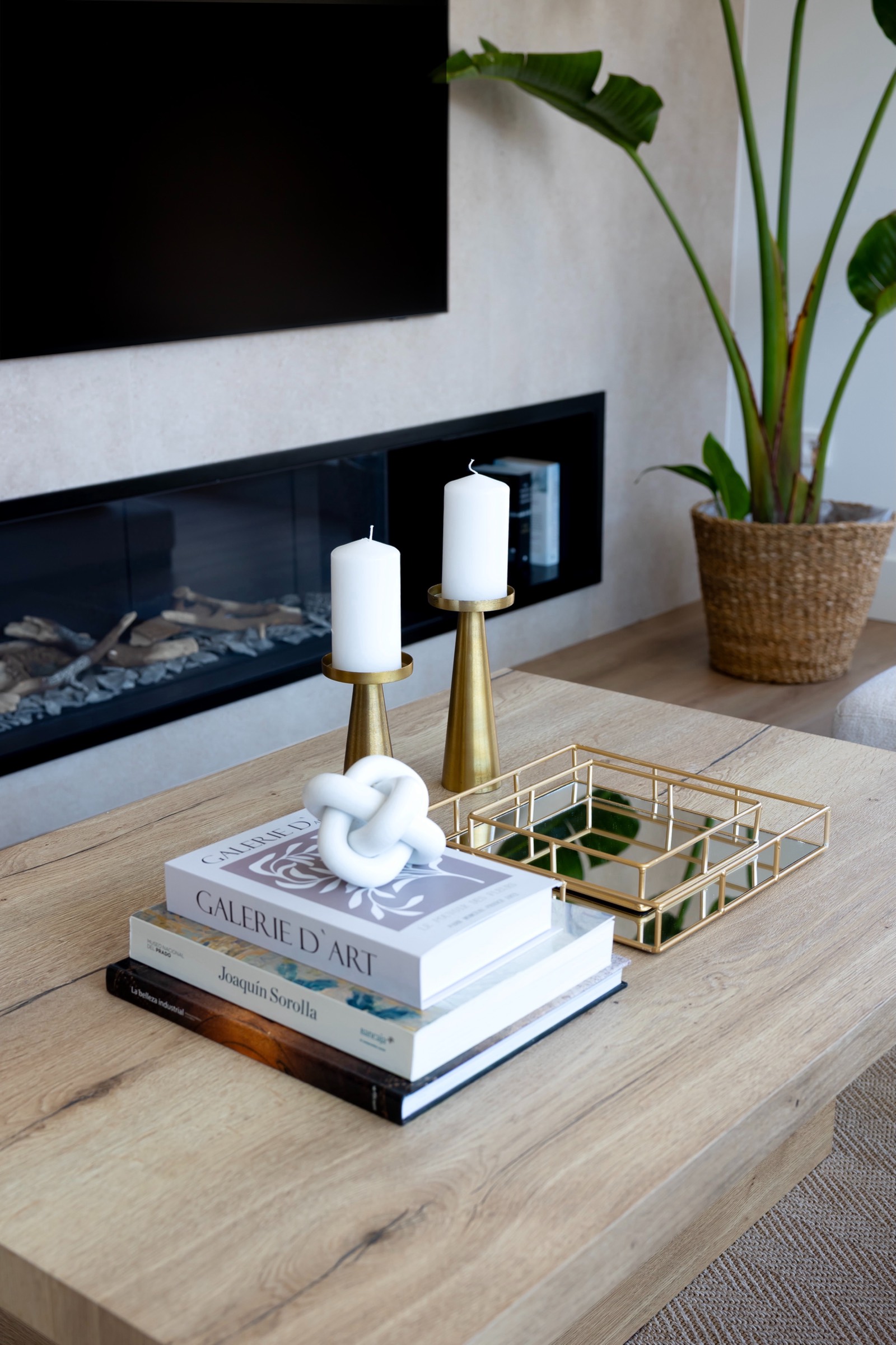 Sala de estar moderna con chimenea, mesa de centro de madera decorada con libros, velas doradas, y una planta en maceta. La decoración incluye elementos en tonos neutros y detalles en dorado.