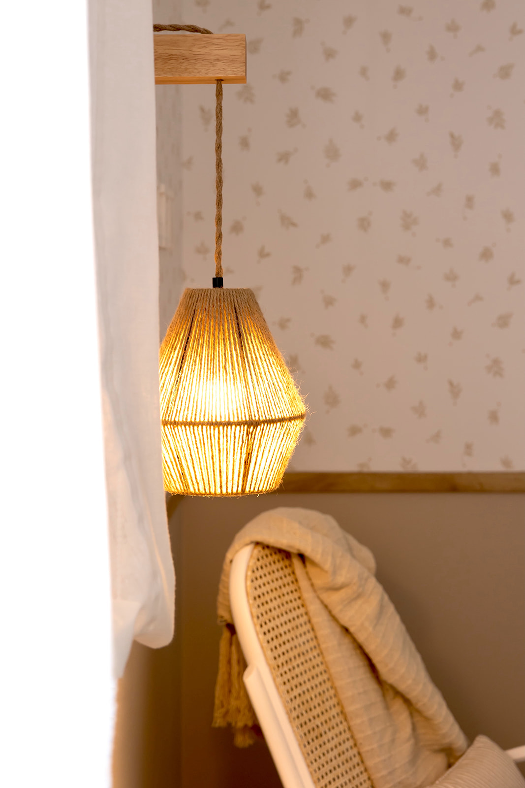 Lámpara colgante de estilo bohemio con pantalla de cuerda trenzada, emitiendo una luz cálida. Está suspendida de un soporte de madera y se encuentra junto a una silla de ratán con un cojín beige.