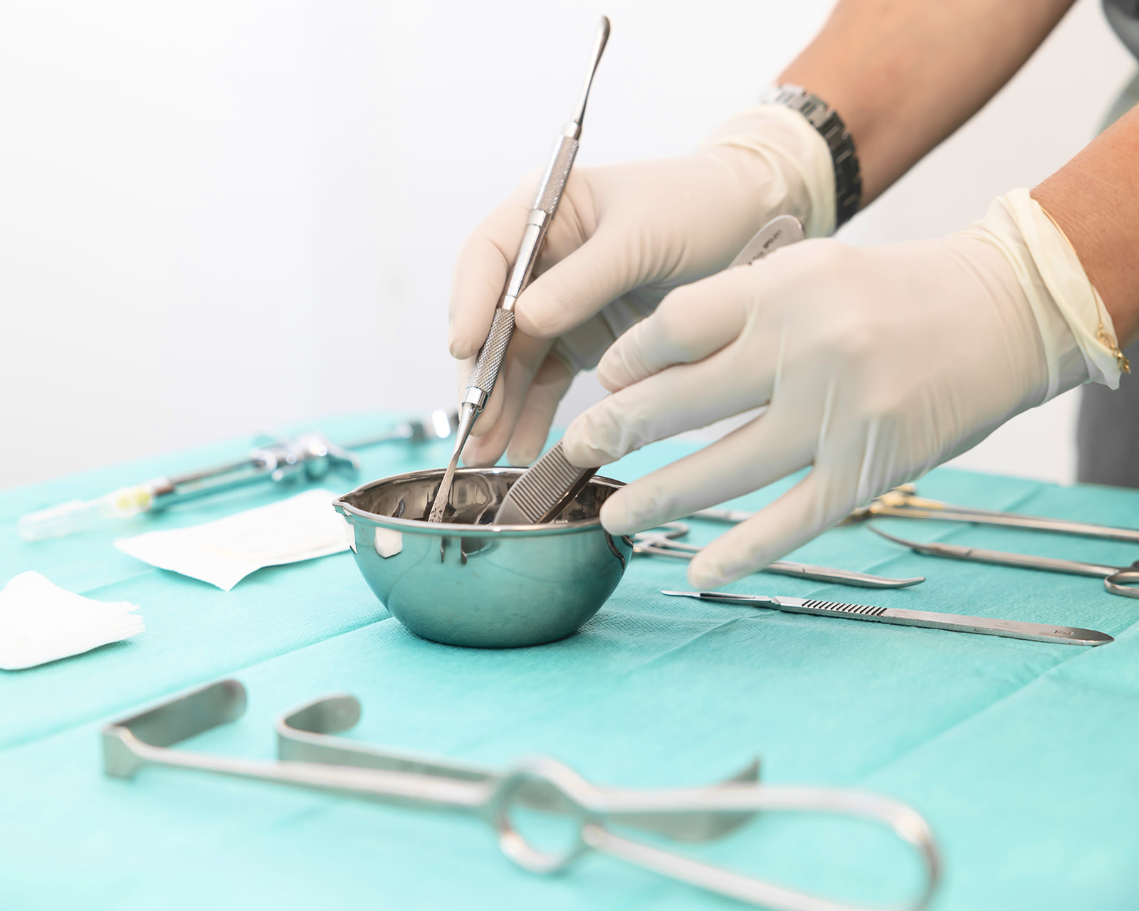 Manos con guantes quirúrgicos preparando instrumental médico en un recipiente metálico sobre una superficie verde. Varios instrumentos quirúrgicos están dispuestos alrededor.